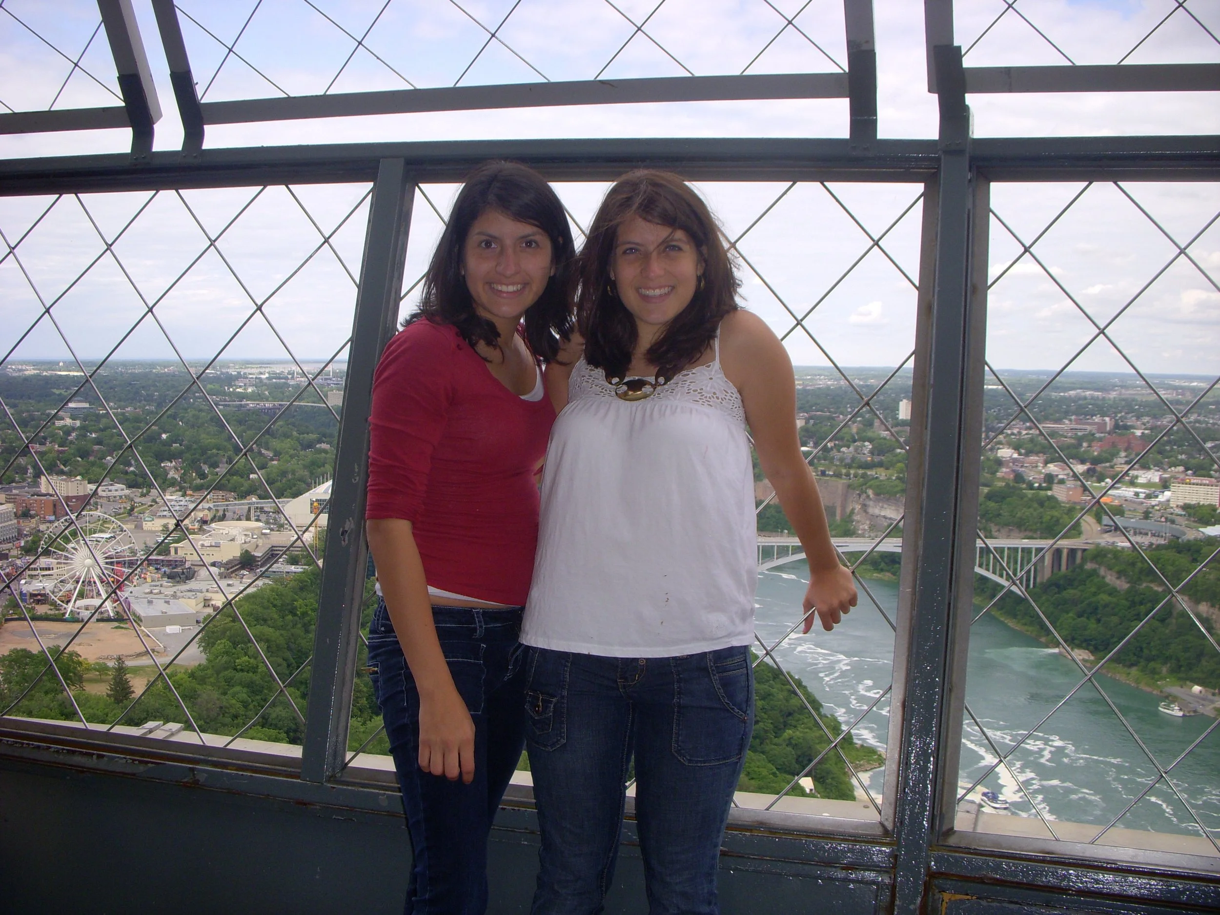 Two women standing on a high observation platform with a cityscape and river in the background, surrounded by metal safety barriers.