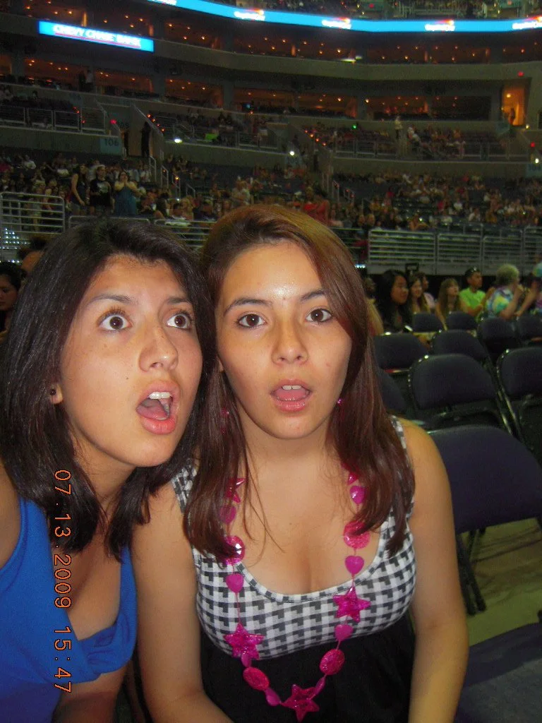 Two young women sitting in a crowded arena or theater with surprised or shocked expressions, one wearing a blue top and the other wearing a checked top with pink necklaces.