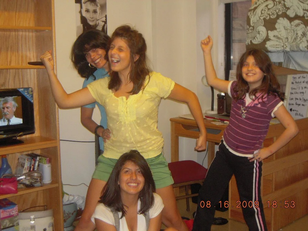 Four young women posing and smiling inside a room, with a TV showing a man on the screen and a poster of a woman on the wall.
