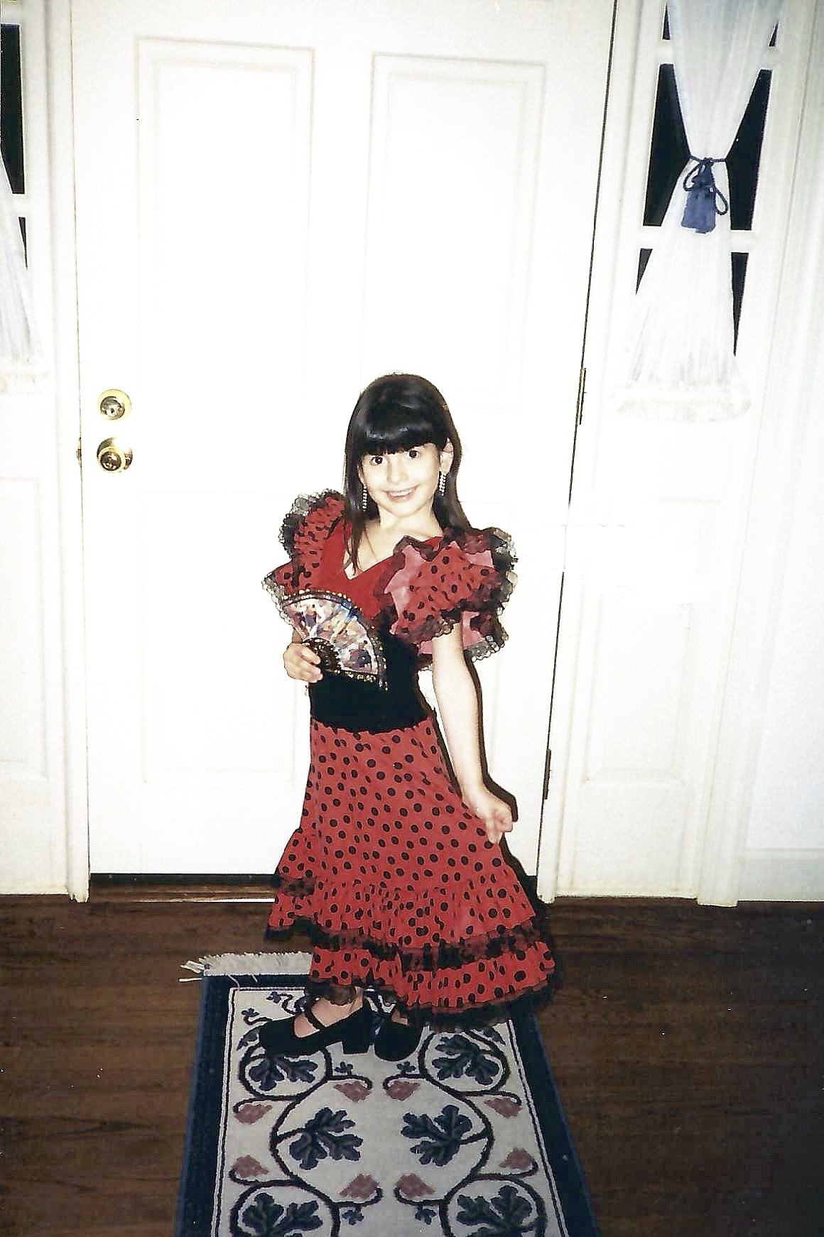 A woman in a red and black flamenco-style dress holding a colorful fan, standing on a decorative rug in front of a white door with black and white wall decorations.