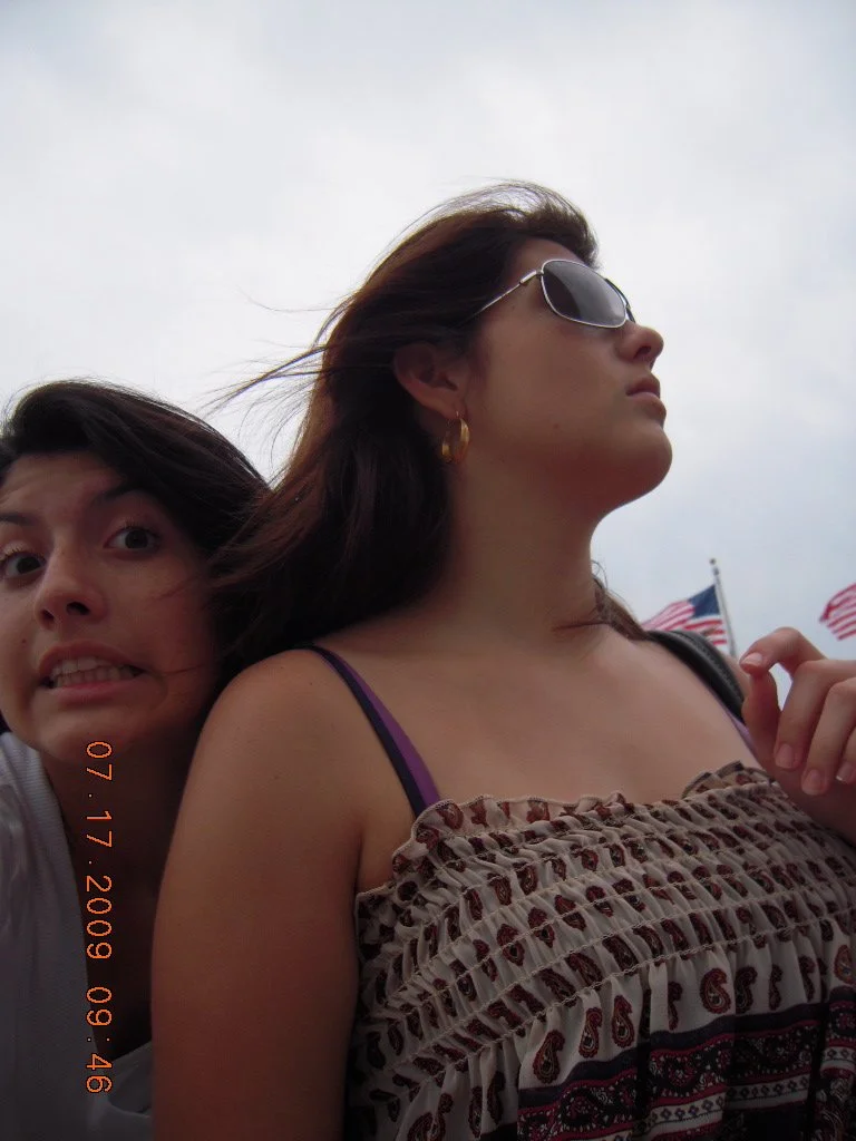 Two women are outdoors, one with long brown hair wearing sunglasses and a patterned dress, the other with dark hair making a funny face, with American flags in the background.