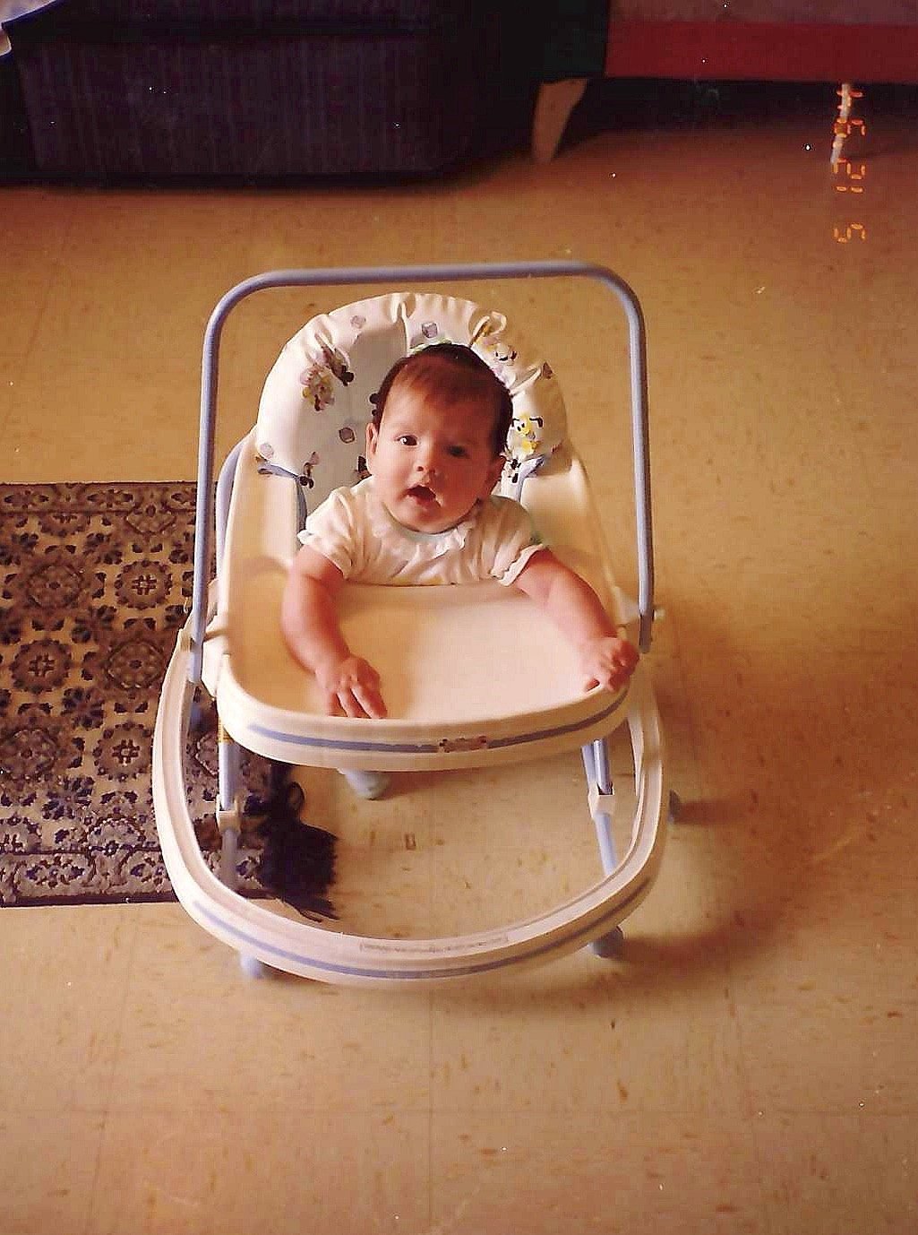 A baby sitting in a white baby walker on a beige floor near a small rug, with a childlike expression, in a room with dark furniture and a warm-colored wall.
