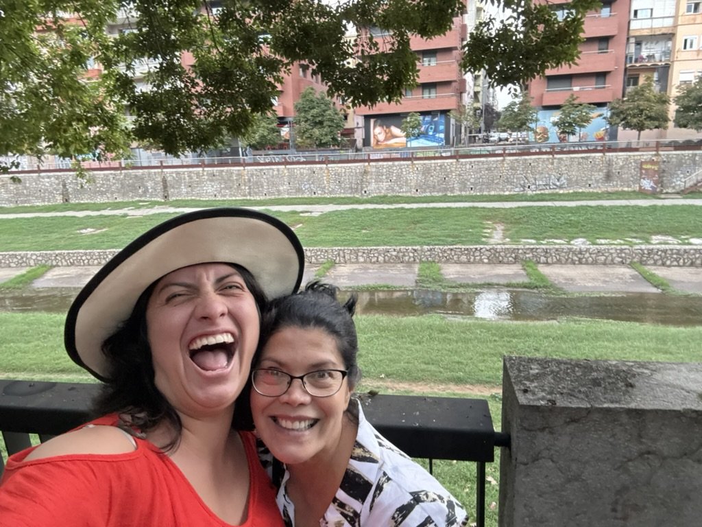 Two women smiling and laughing together outdoors in a park, with a river and buildings in the background.