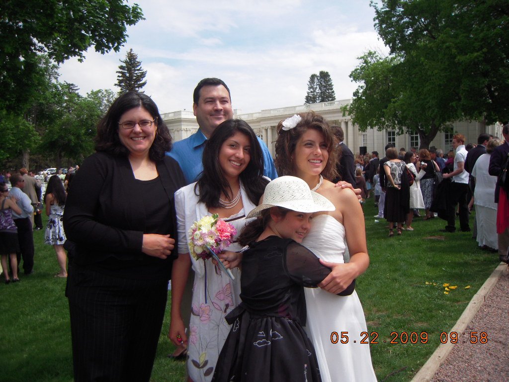 Group of five people, including a bride in a white wedding dress with a flower in her hair, standing outdoors on a grassy area during a wedding celebration. Multiple guests are visible in the background, with some gathered near a white building, tree