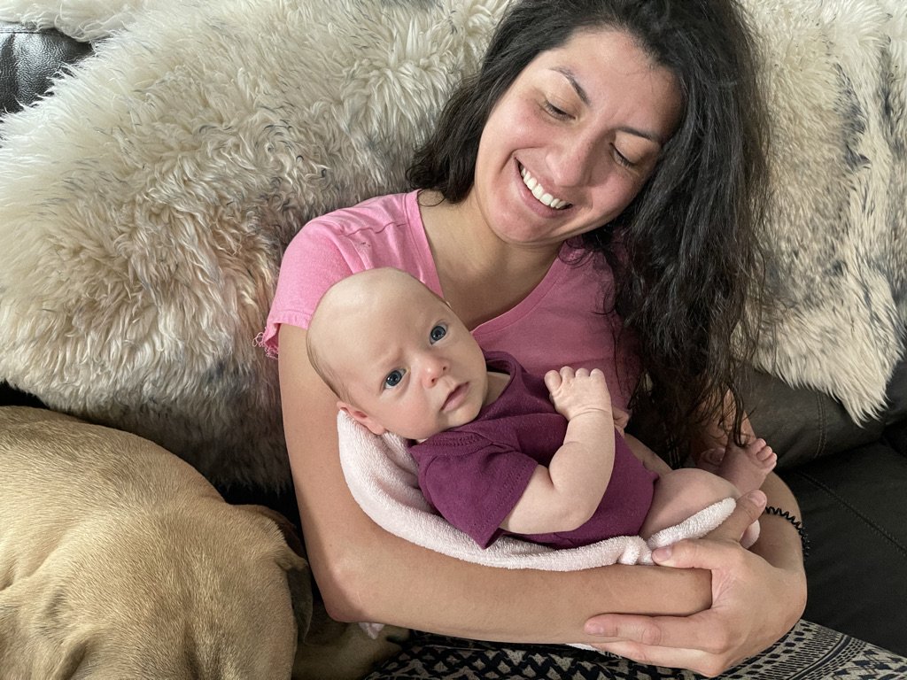 A woman with dark hair smiling and holding a baby with blue eyes, both sitting on a couch with fluffy pillows. A dog is lying beside them.