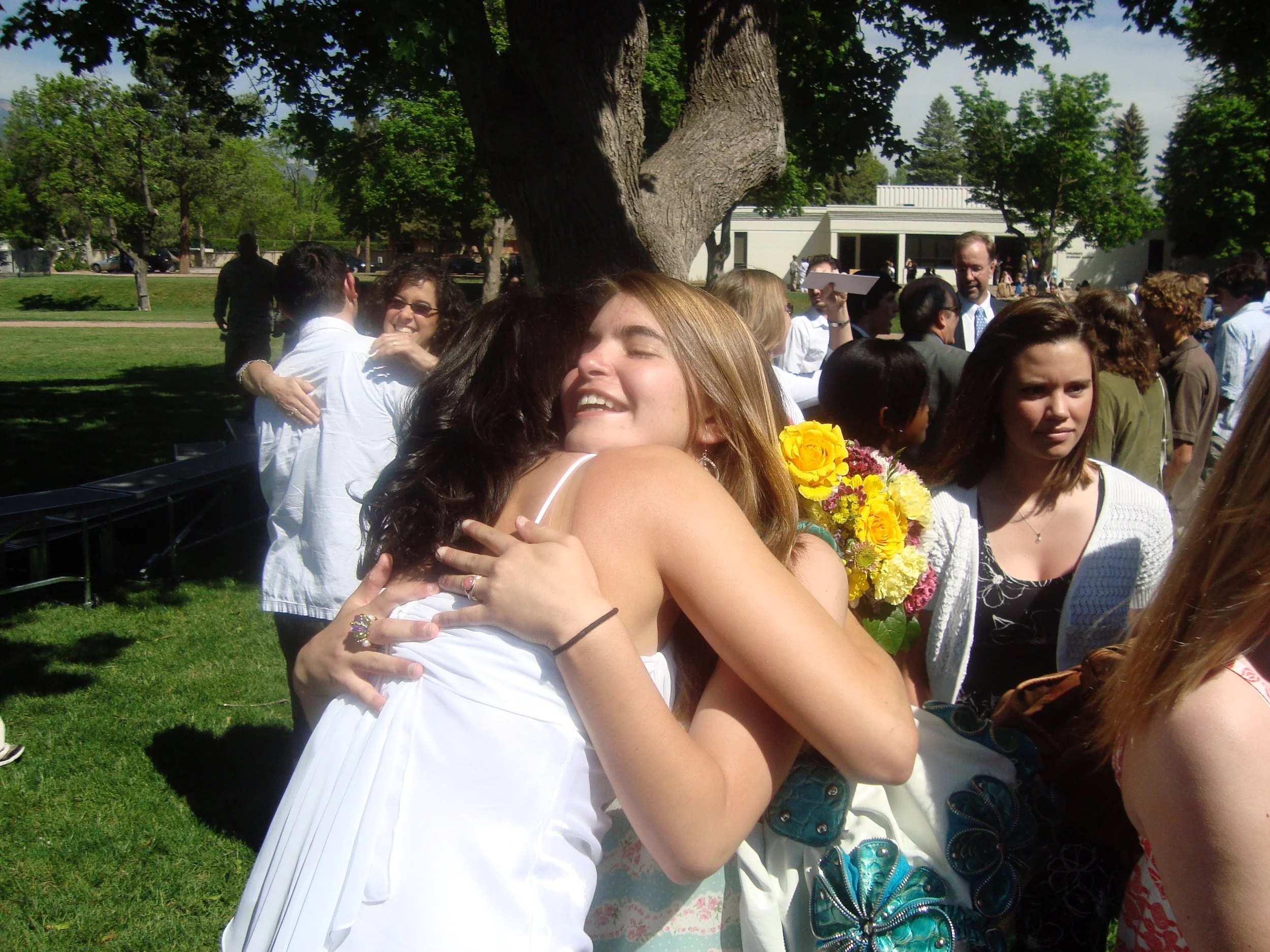 Group of people hugging and smiling outdoors under a large tree at a sunny event, with some holding bouquets of flowers.