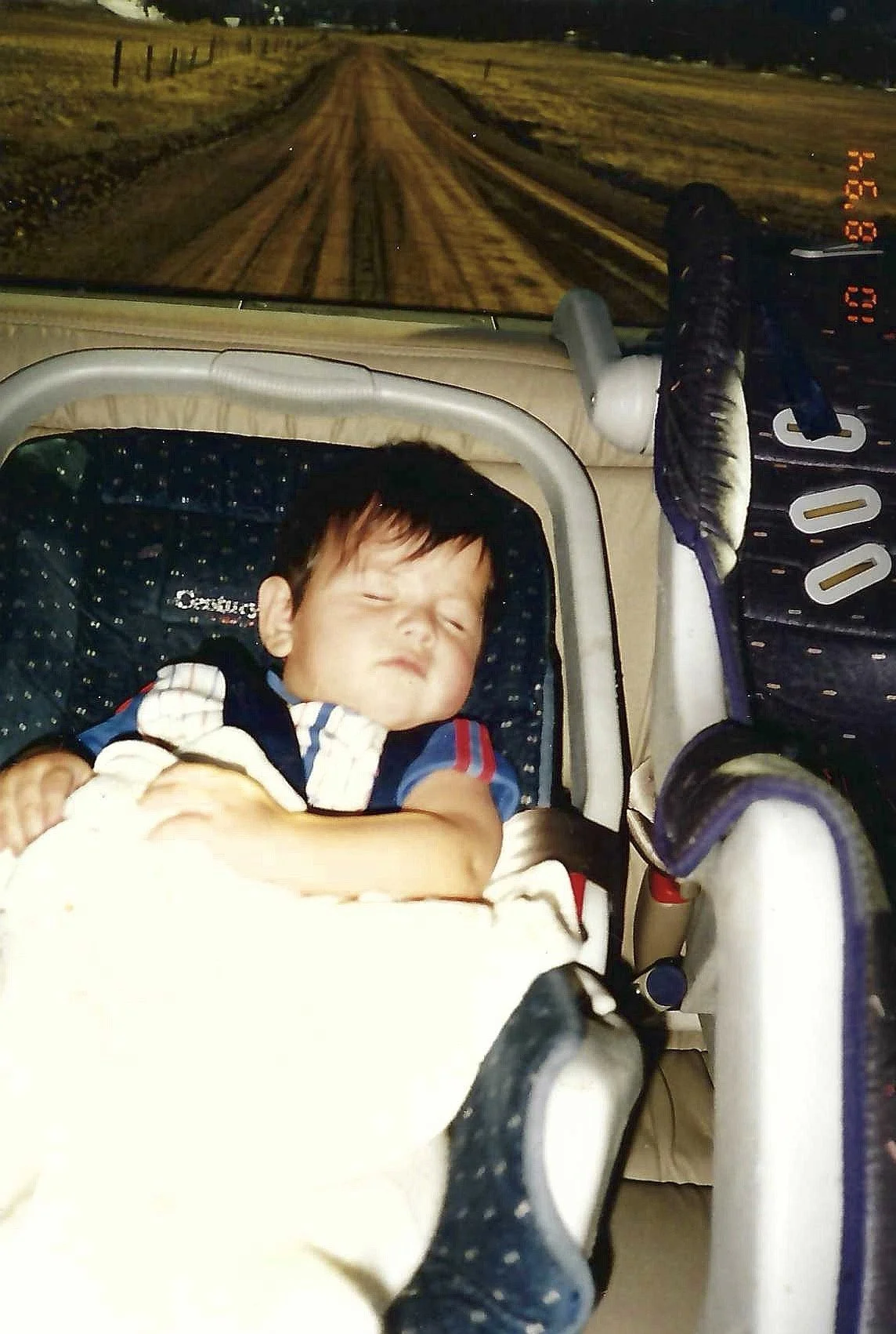 A young boy sleeping in a car seat inside a vehicle, with a view of a dirt road and fields visible through the windshield at night.