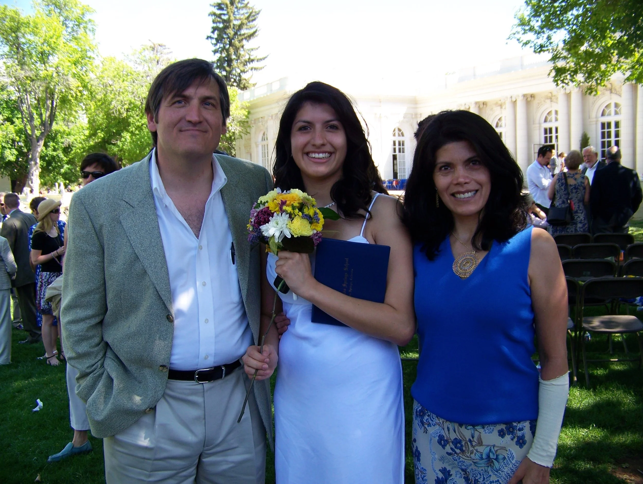 Three people smiling at an outdoor event, with a woman holding a bouquet of flowers. The background shows trees and a white building with large windows.