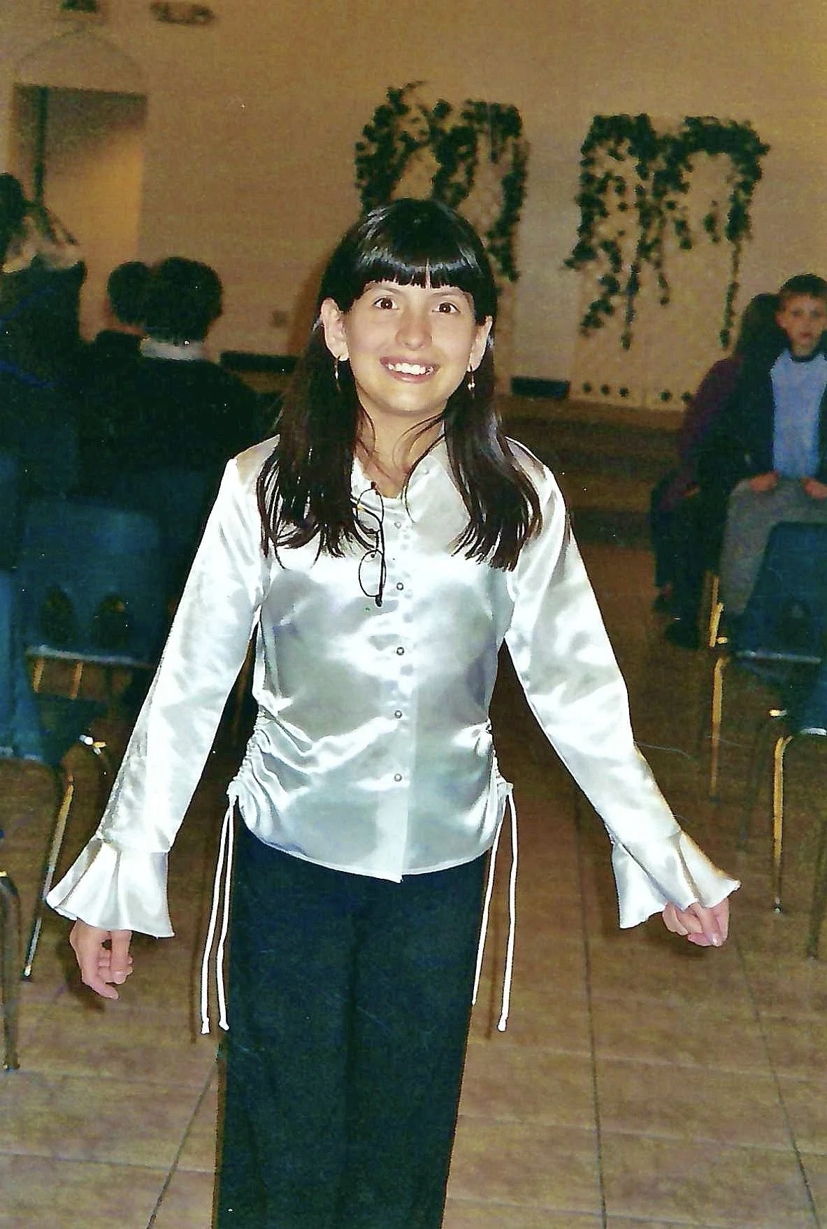 A woman in a shiny satin blouse with ruffled bell sleeves standing in a room with chairs, other people, and wall decorations.