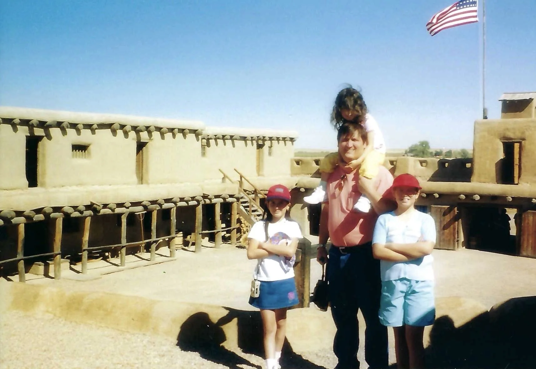 Four children and one adult standing in front of a historic adobe-style building with an American flag, with blue sky and open landscape in the background.