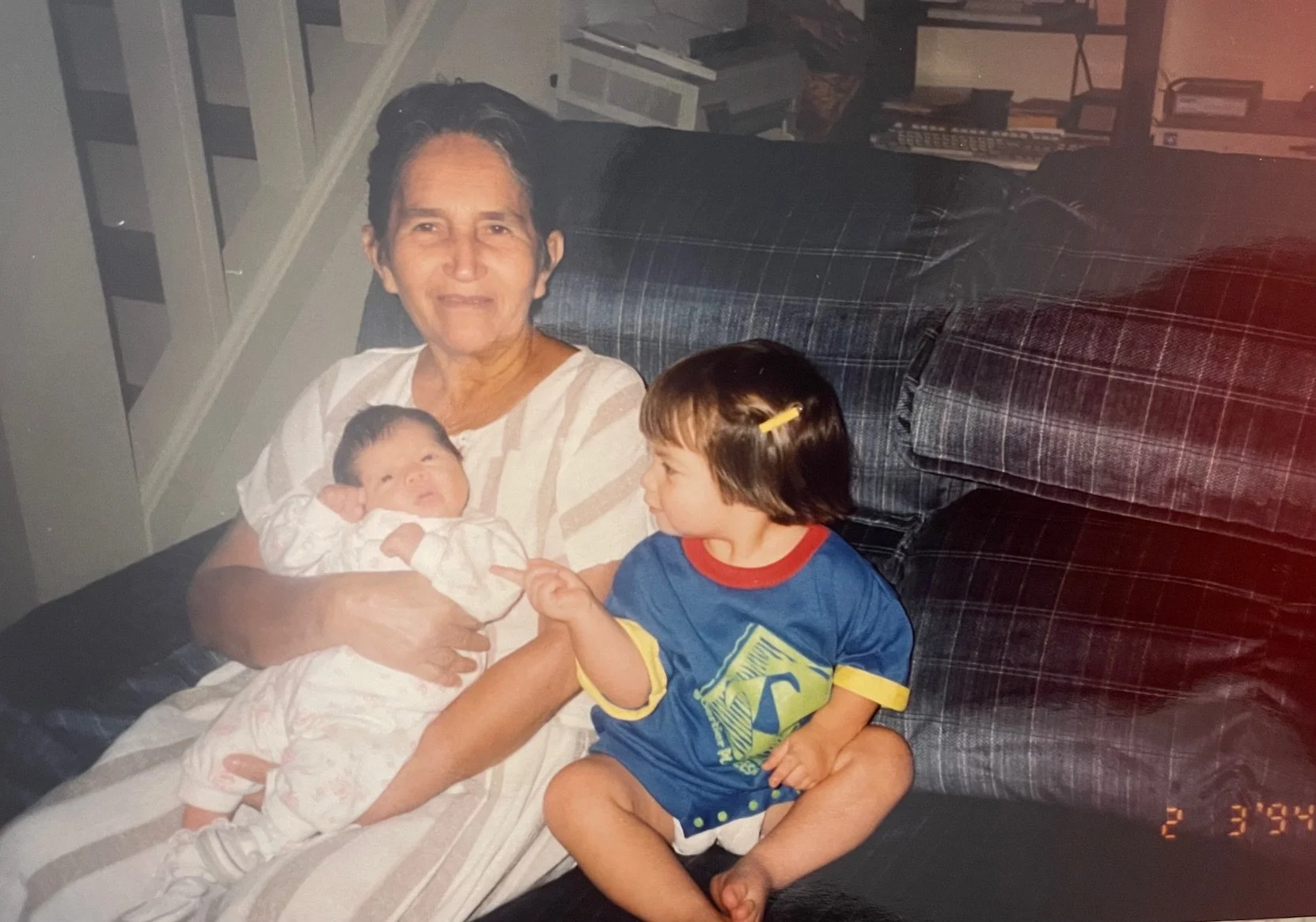 An elderly woman sitting on a plaid sofa holding a newborn baby in her arms, and a young girl with short brown hair, wearing a blue T-shirt with a yellow graphic and a yellow hair clip, sitting on her lap and pointing at the baby, in a living room wi