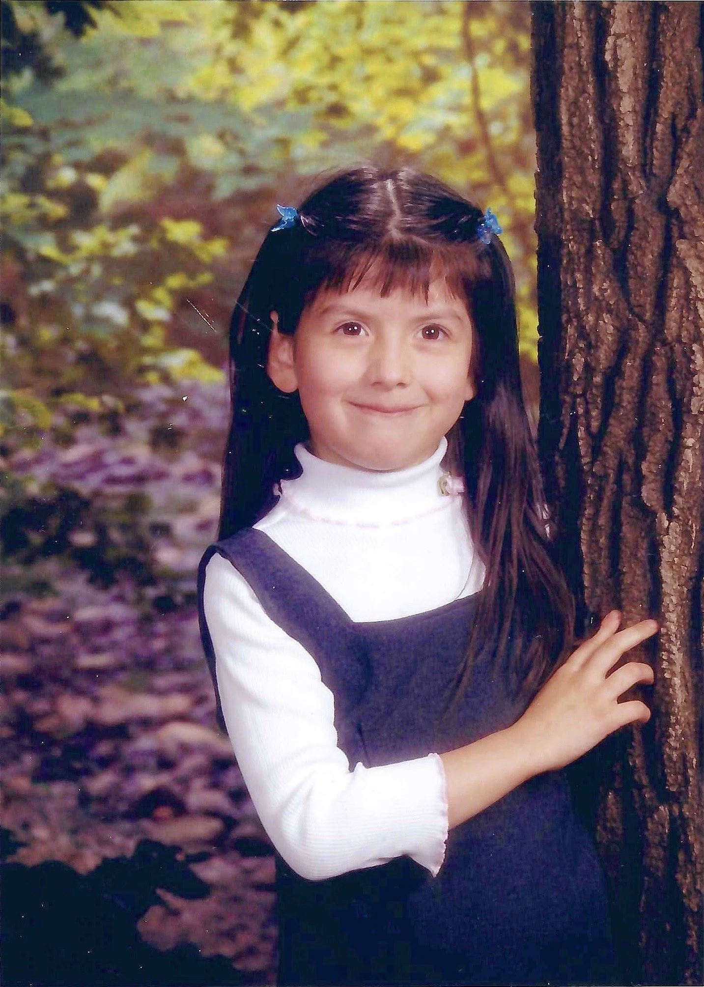 A young girl with dark hair and blue hair clips, wearing a white turtleneck and a navy dress, standing outdoors next to a tree with autumn leaves in the background.