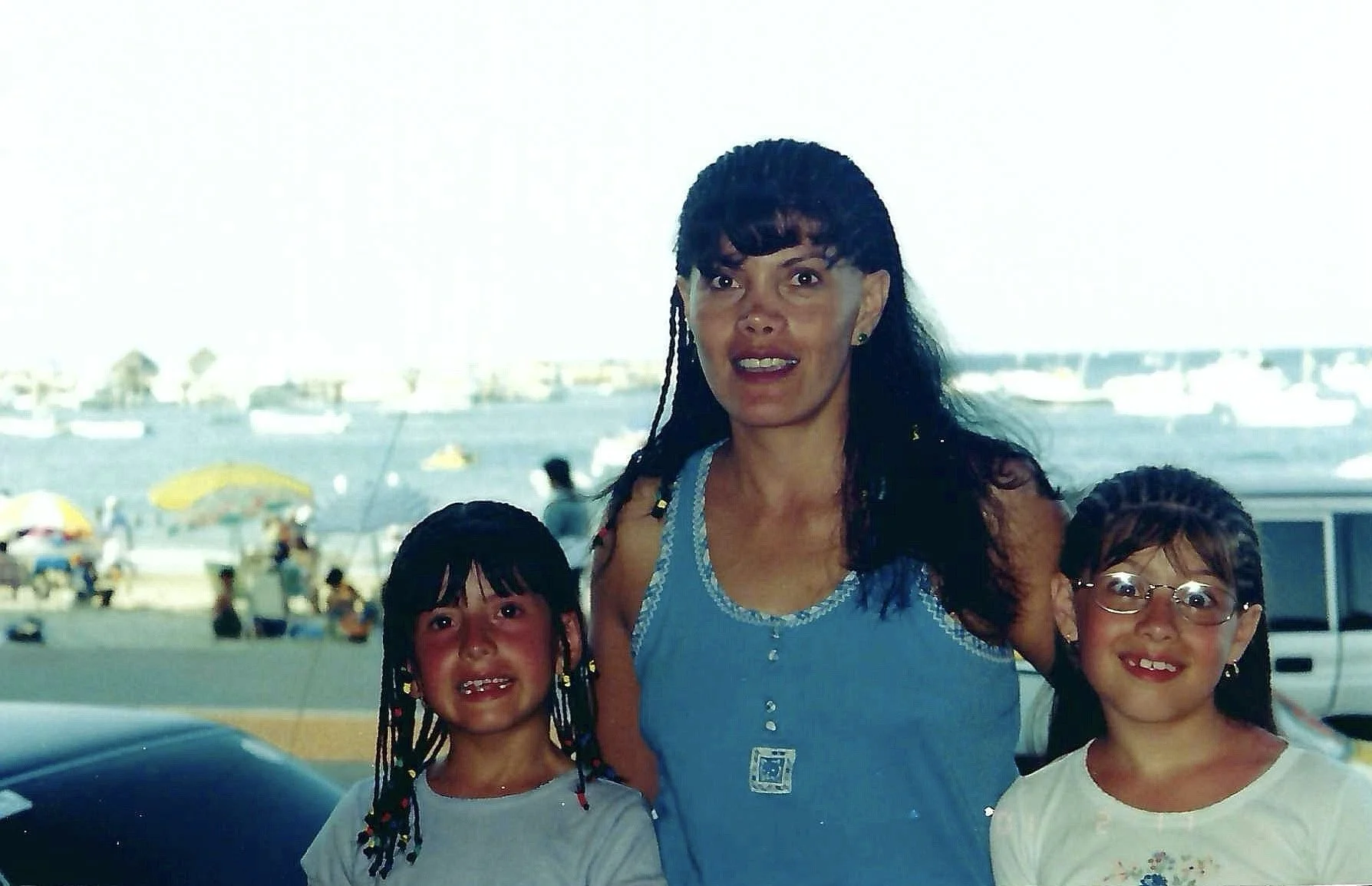A woman and two children posing together at a waterfront, with boats and people on the beach in the background.