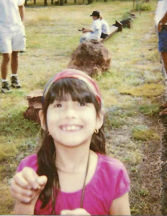 A young girl smiling and wearing a pink shirt and headband, with people sitting and standing on logs in an outdoor park setting in the background.