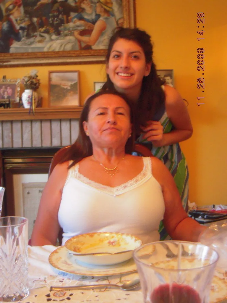 A woman and a young girl smiling at a dining table inside a home, with a yellow wall, framed pictures, and a painting behind them.