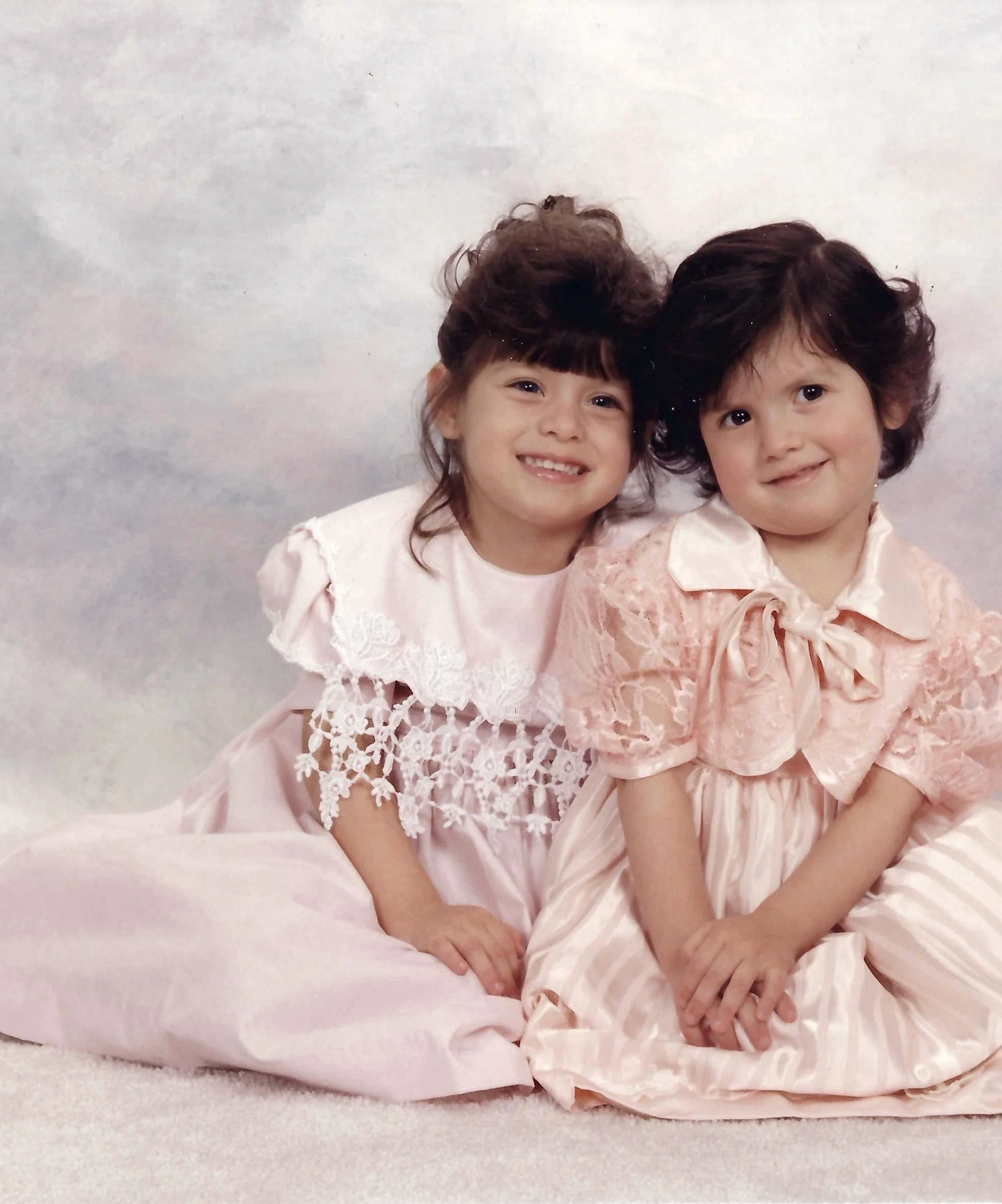 Two young children, a girl and a boy, sitting close together on a soft pink blanket, both smiling and dressed in pink lace dresses with puffed sleeves and lace details, against a light-colored background.