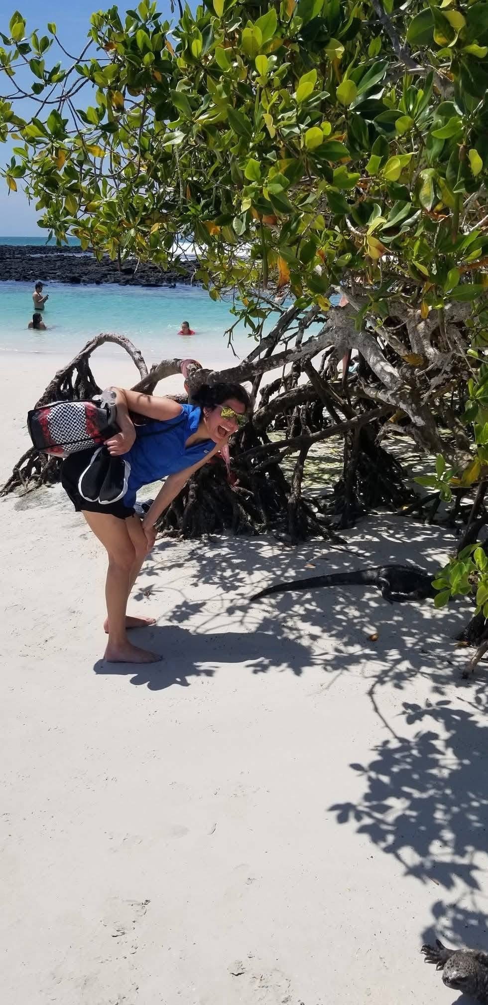 A smiling woman with sunglasses, a blue shirt, and black shorts, crouching on a sandy beach. She is carrying a checkered bag and posing near a large mangrove tree with twisted roots and green leaves. In the background, people are swimming in clear tu