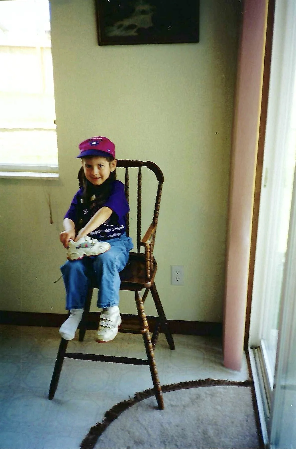 A young girl wearing a pink cap, purple shirt, and blue jeans, sitting on a wooden chair that is stacked on top of another chair, indoors near a window and a pink wall.