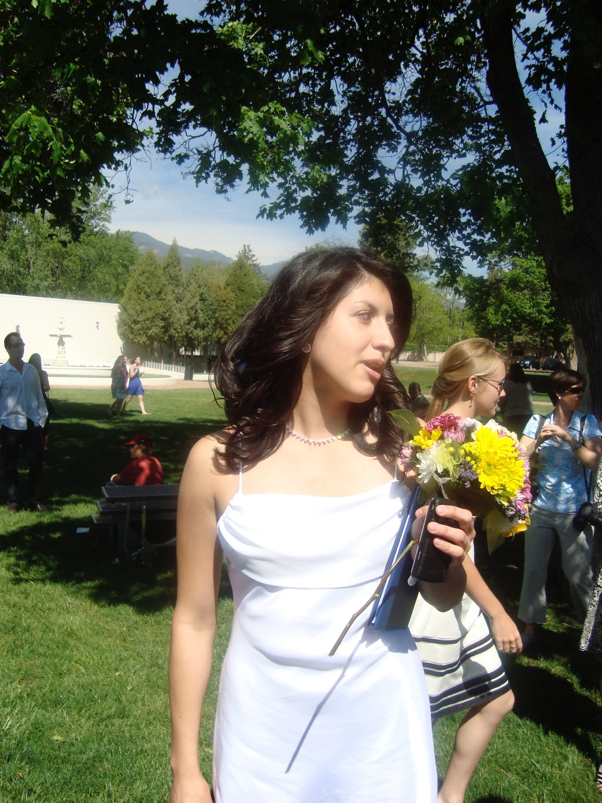 A young woman with long dark hair wearing a white dress, holding a bouquet of colorful flowers, standing outdoors under a large tree with other people in the background.