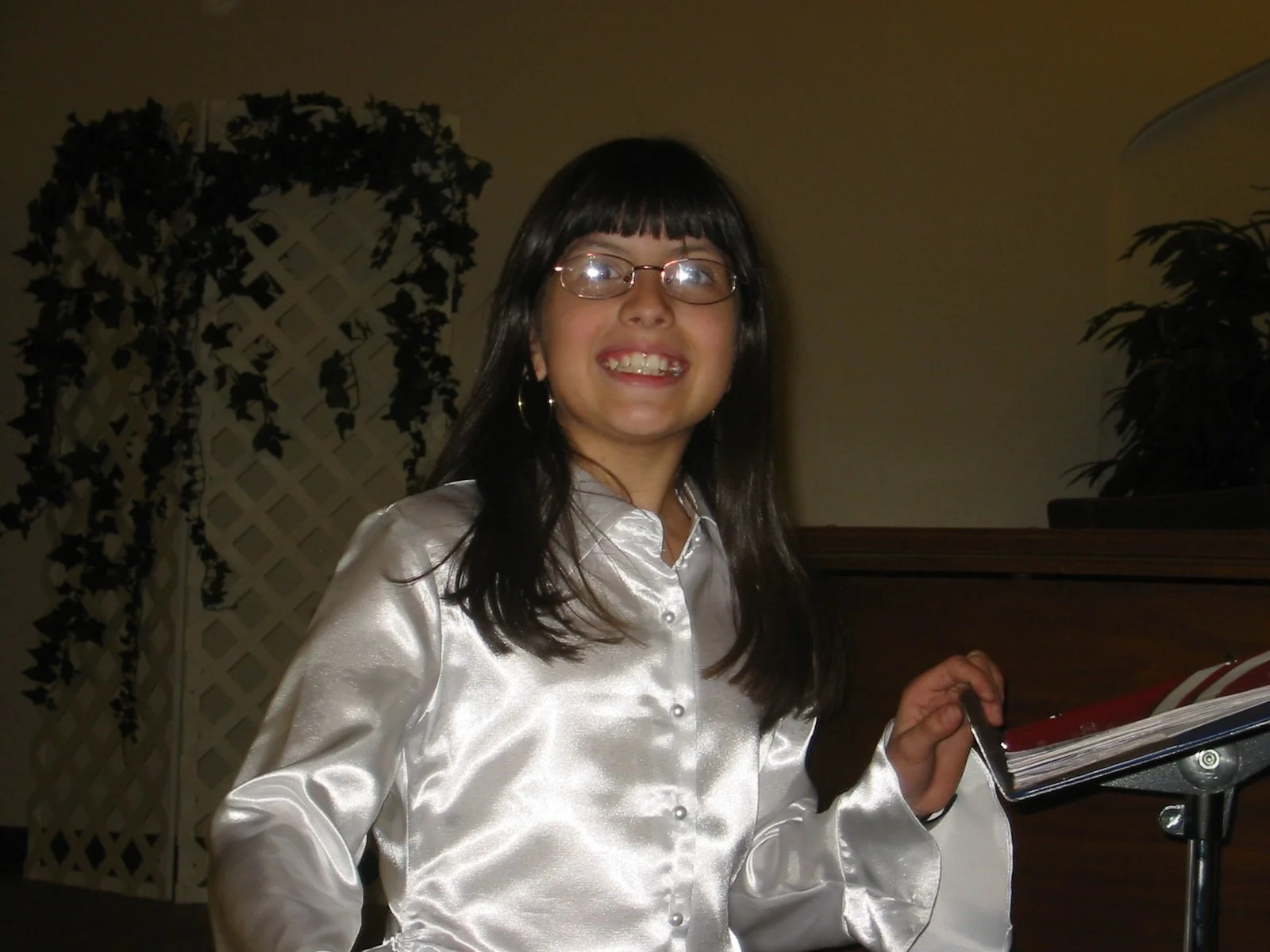 Smiling woman with dark hair, glasses, and hoop earrings, wearing a shiny white blouse, holding a book or binder, standing indoors with plants in the background.
