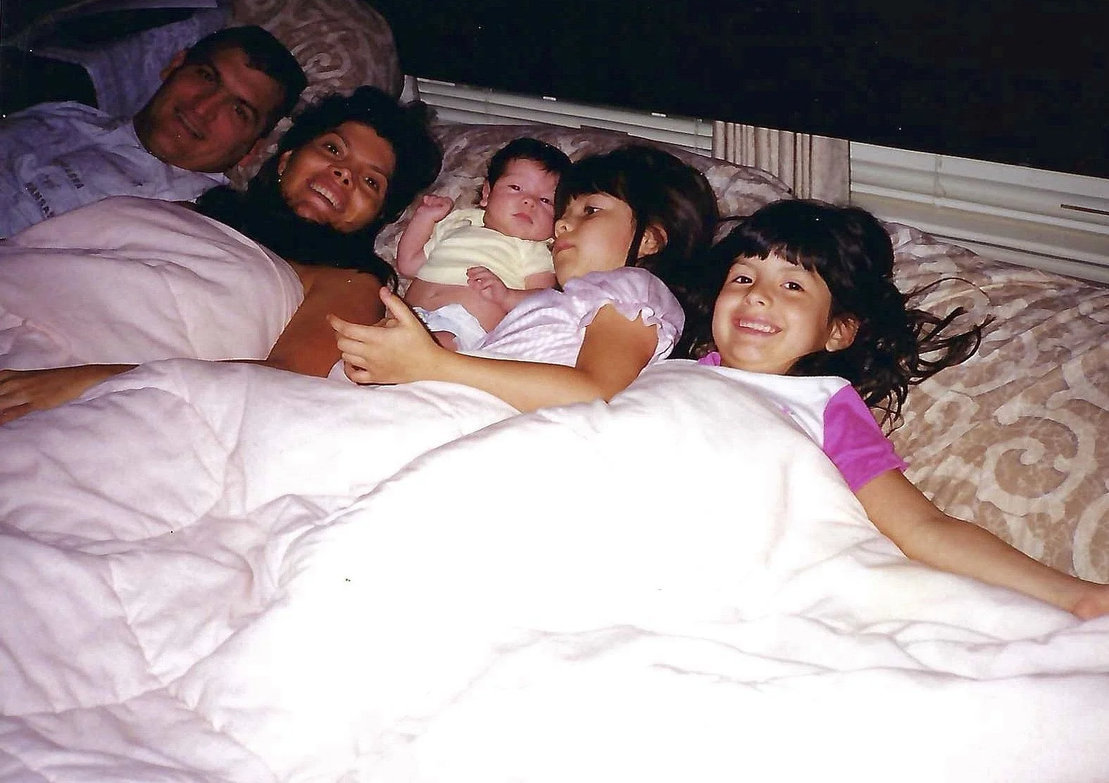 A family of five lying on a bed together, smiling and relaxing. The father and mother are at the head of the bed with three children, including a baby, lying between them. The children are smiling, and the overall mood is happy and cozy.