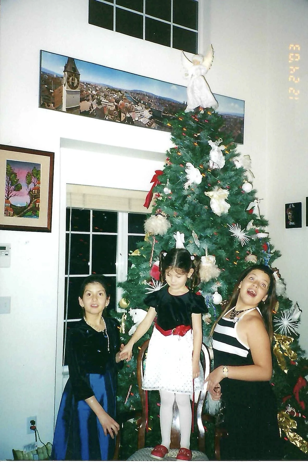 Three young girls standing in front of a decorated Christmas tree with a star on top, inside a house with wall art and a window. They are holding hands and smiling.