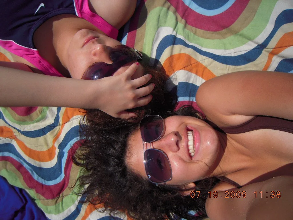 Two women lying on a colorful striped beach towel, both wearing sunglasses and smiling at the camera. One woman has curly dark hair and the other has lighter-colored hair.