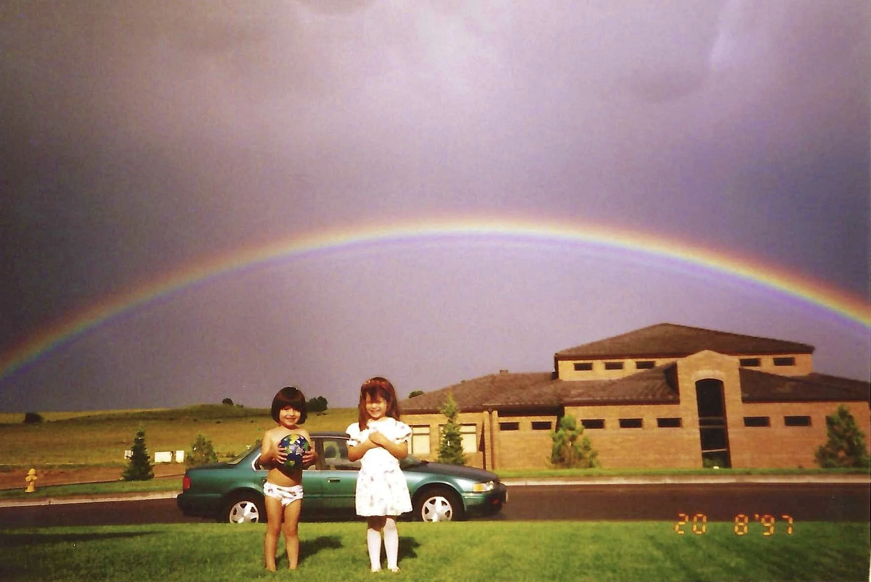 Two young girls standing on grass in front of a vintage green car, with a large house in the background and a rainbow in a rainy sky above, taken on August 20, 1993.