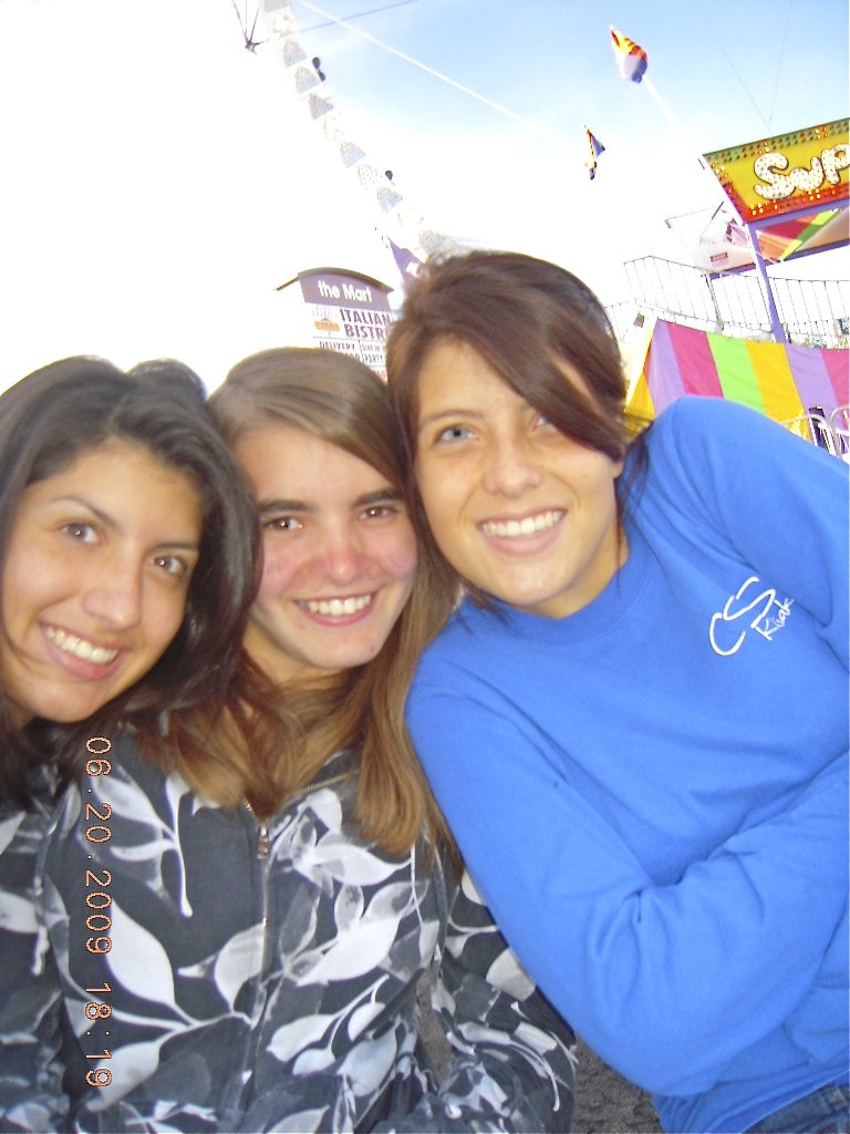 Three young women smiling and posing for a photo at a fair or carnival with booths, flags, and a ride in the background.