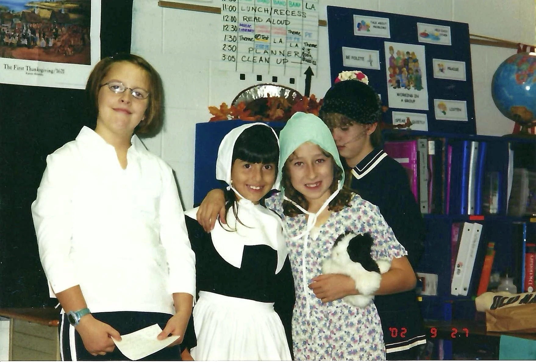 Four young girls in a classroom, dressed in costumes, one holding a black and white rabbit. The background includes educational posters, a bookshelf with binders, and a calendar. The girls are smiling and appear to be participating in a school activi
