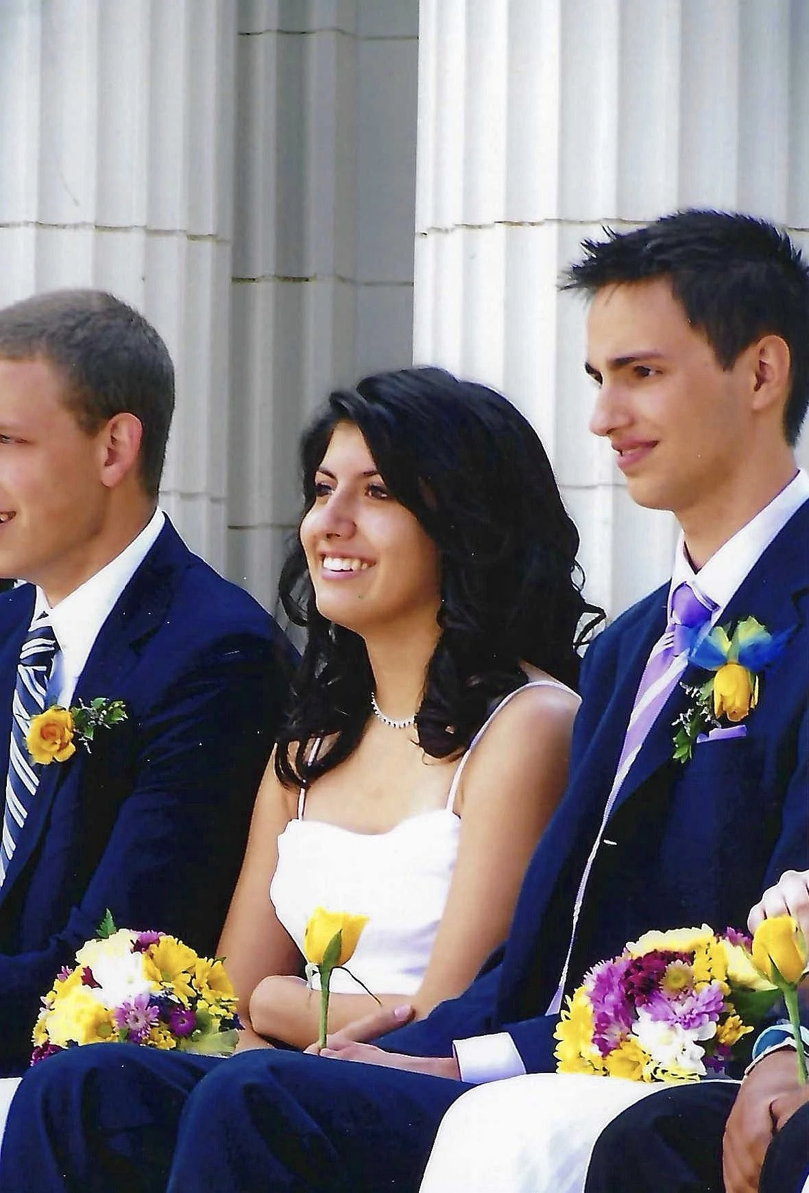 Young woman smiling, dressed in white, sitting with two young men in formal suits, holding colorful bouquets of flowers, against a white architectural background.