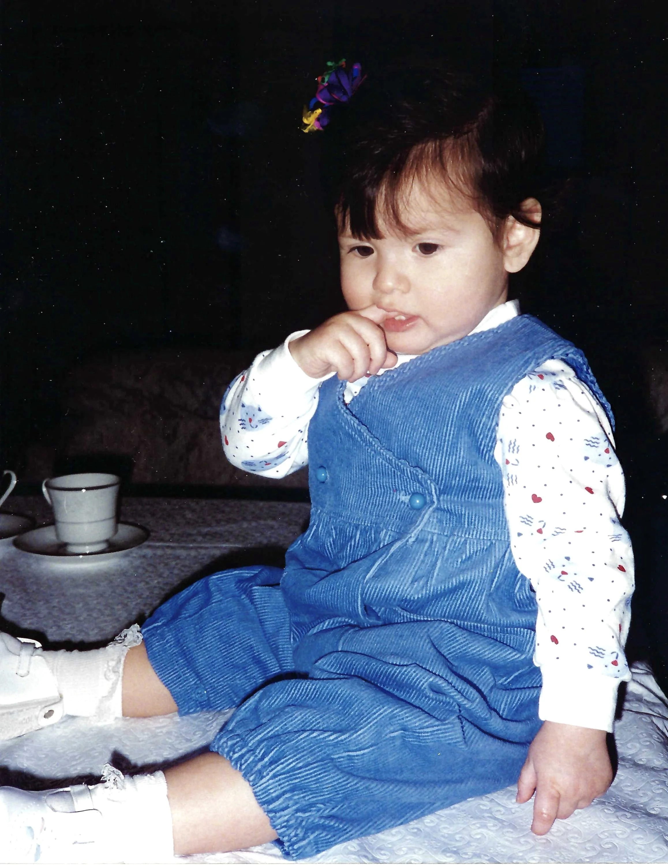 Young child with brown hair wearing a blue overalls, white shirt with red and blue patterns, and white sneakers, sitting on a textured surface near a cup and saucer, with a dark background.
