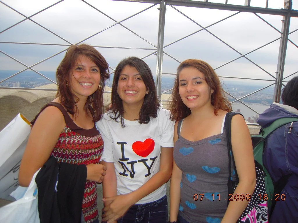 Three young women smiling and standing close together outdoors on a cloudy day with a city view in the background.