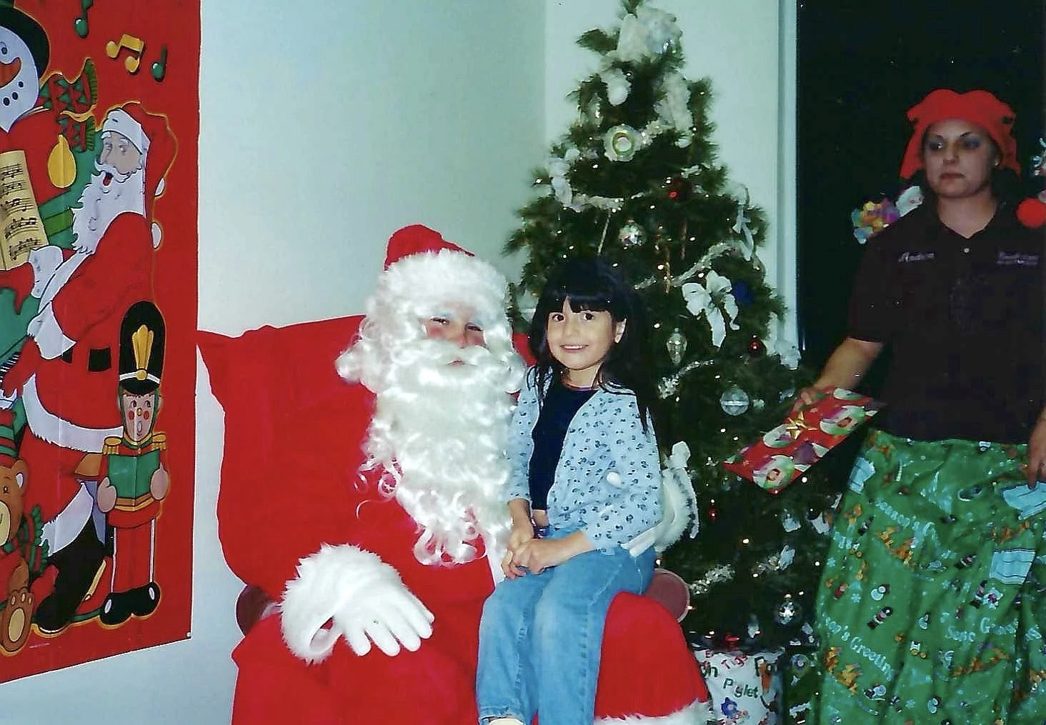 A young girl sitting on Santa Claus's lap during Christmas, with a decorated Christmas tree in the background and a woman holding wrapped presents nearby.