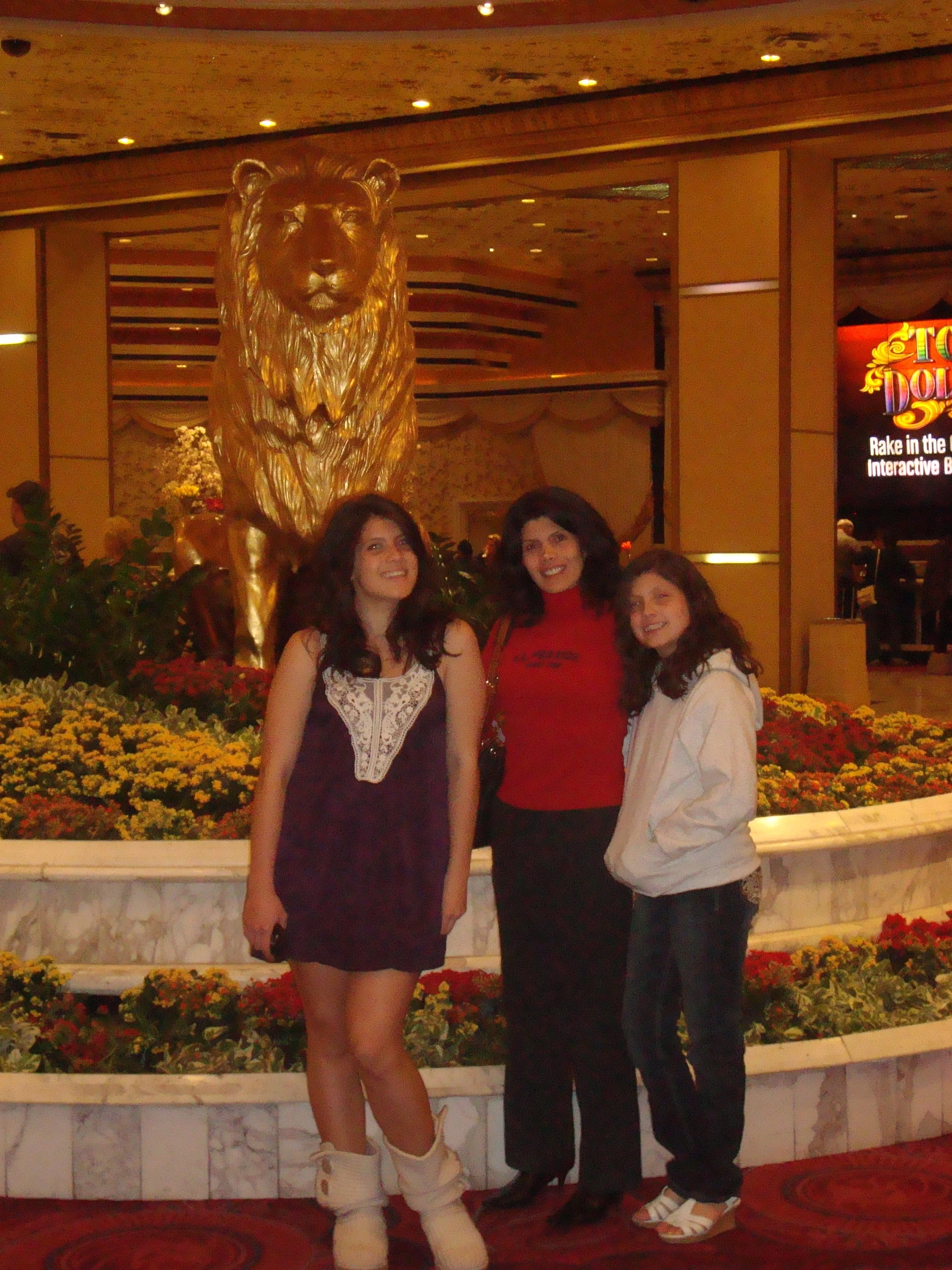 Three women posing in front of a large golden lion sculpture with a flower arrangement background at an indoor event.