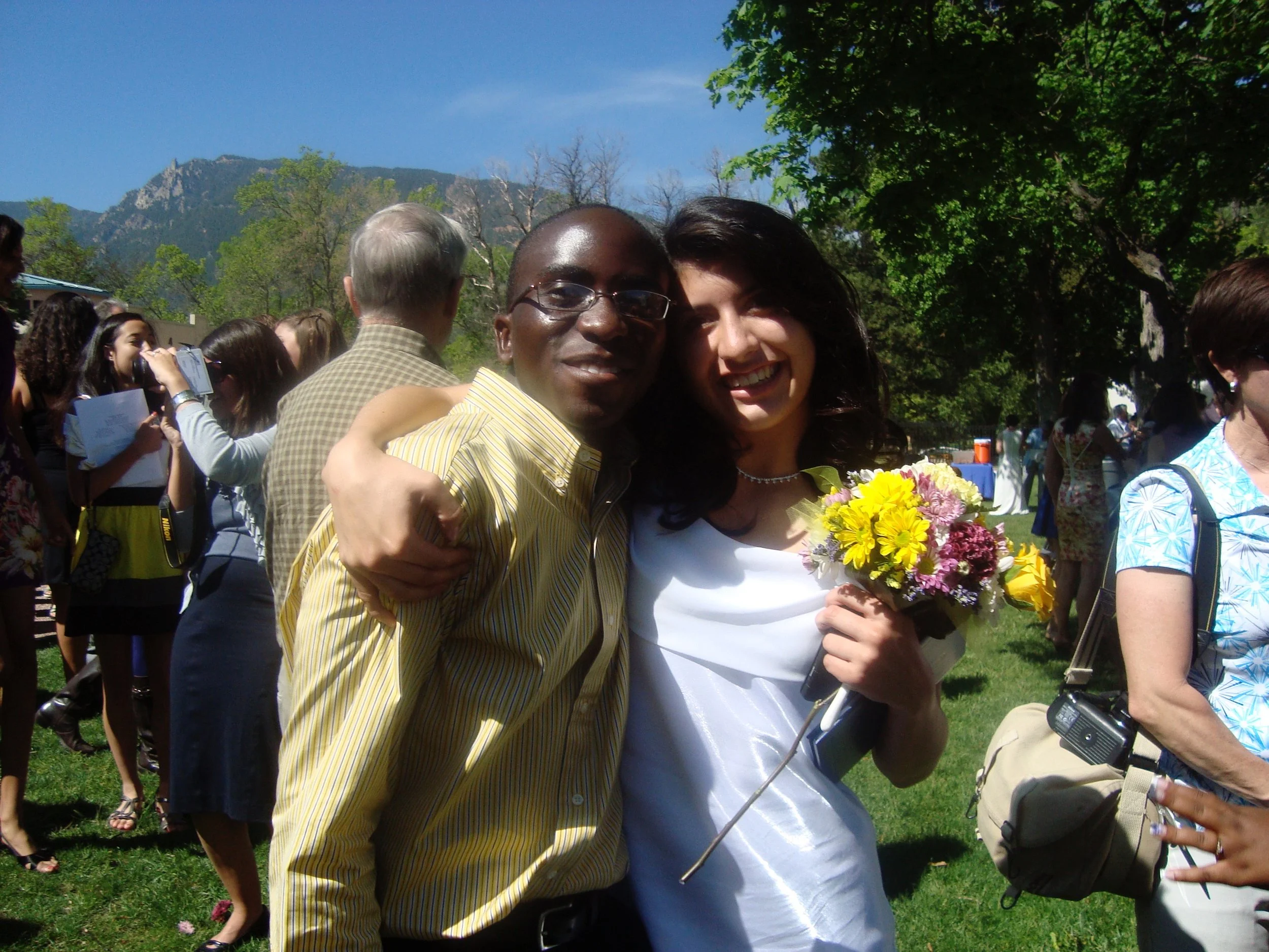 Two people, a man and a woman, are embracing and smiling at an outdoor event. The man is wearing glasses and a yellow striped shirt, and the woman is holding a bouquet of yellow and purple flowers. The background shows a group of people, trees, and m