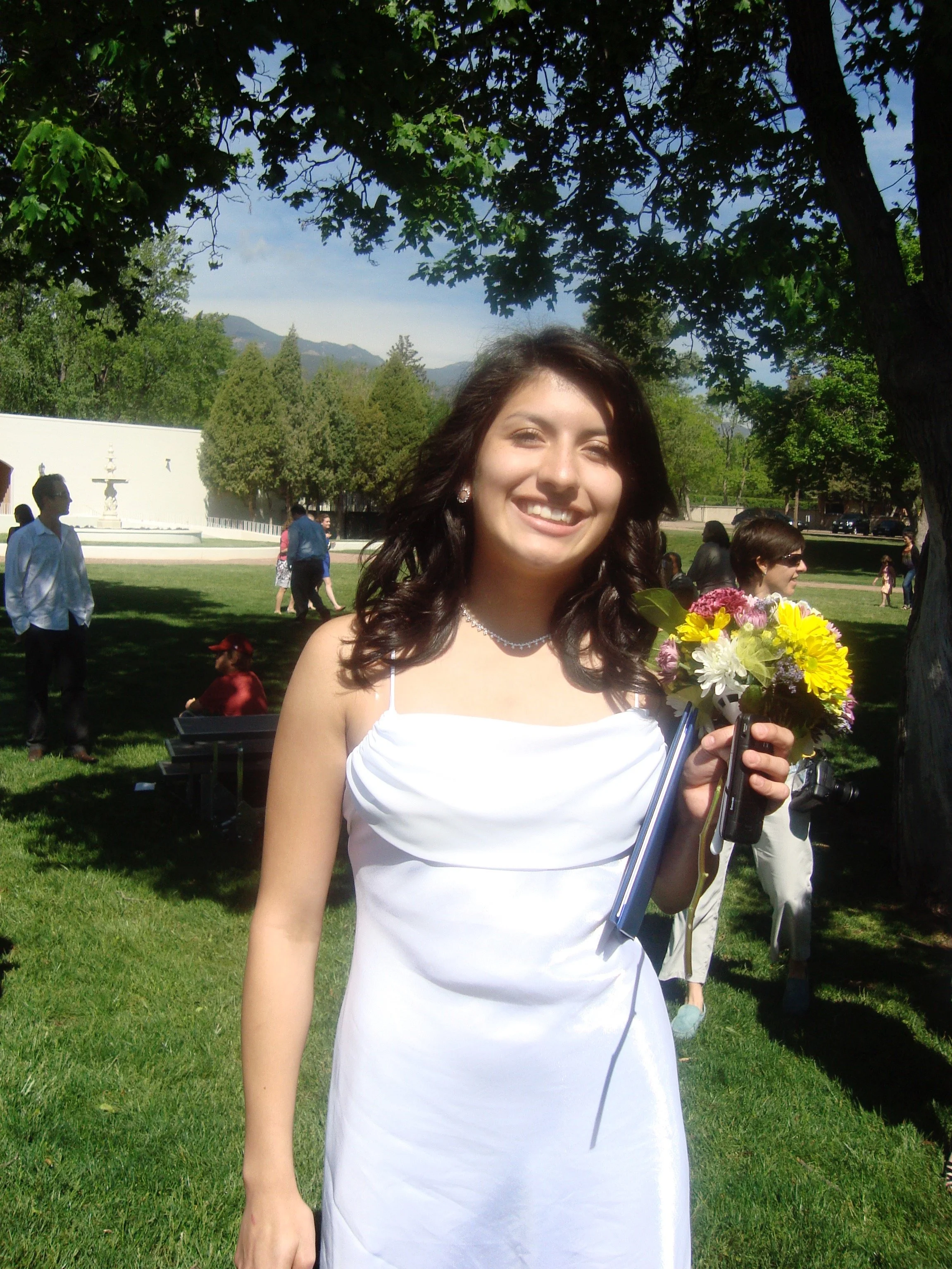 A young woman in a white dress smiling and holding a bouquet of colorful flowers outdoors in a park with other people and trees in the background.