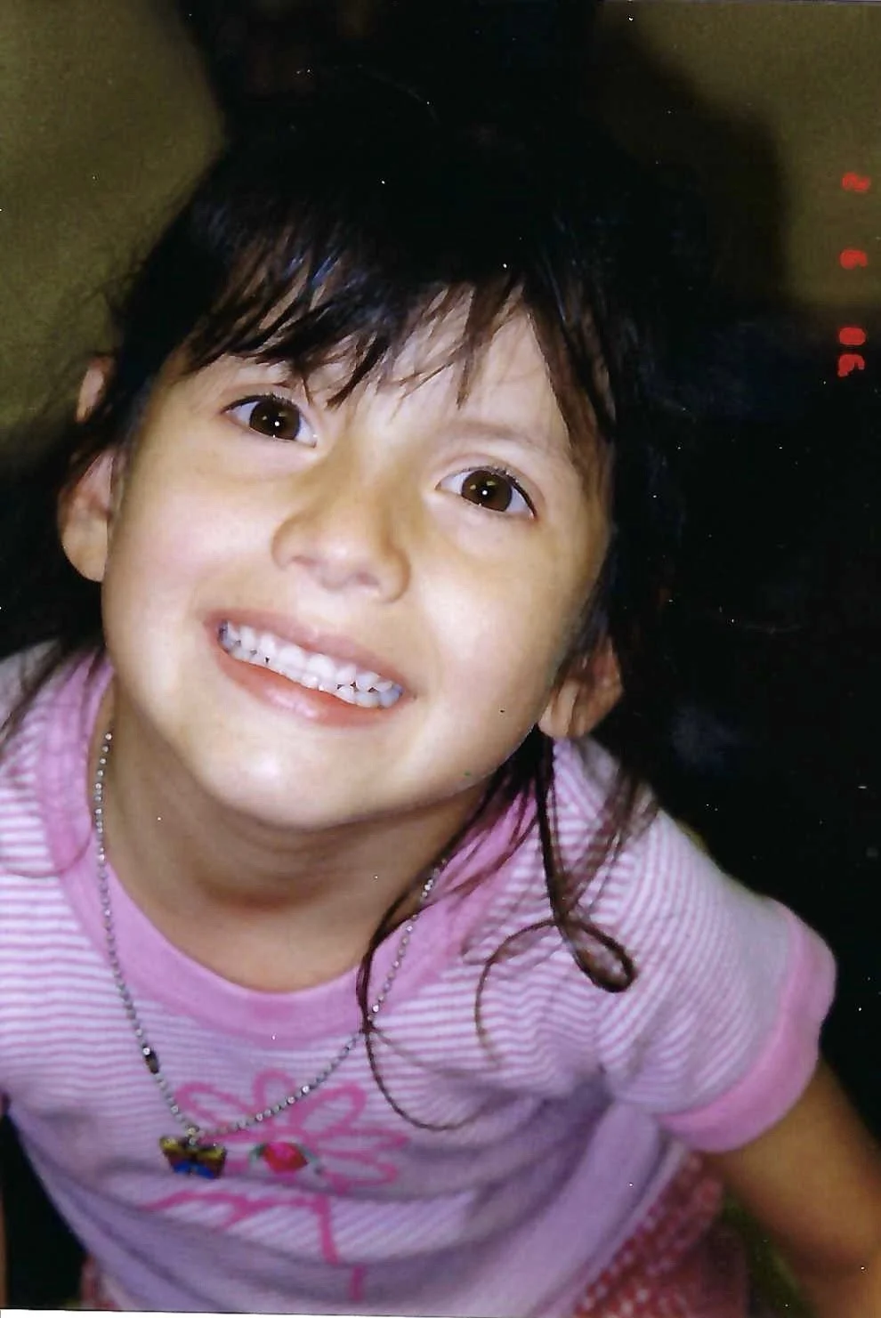 Close-up of a smiling young girl with dark hair, wearing a pink striped shirt and a necklace, looking up at the camera.