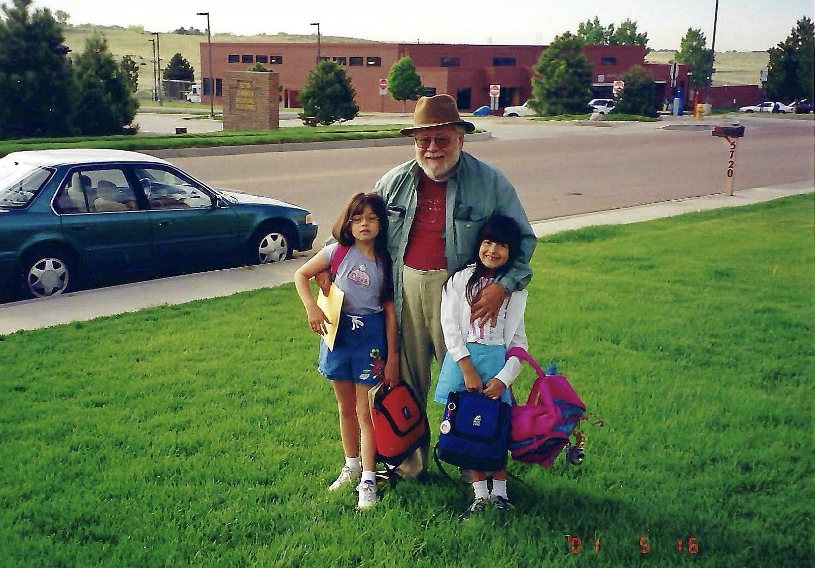 An elderly man with white hair, beard, glasses, wearing a brown hat, red shirt, and gray jacket, standing on green grass with two young girls, all smiling. The girls are holding backpacks and standing in front of him. There are cars parked on the str