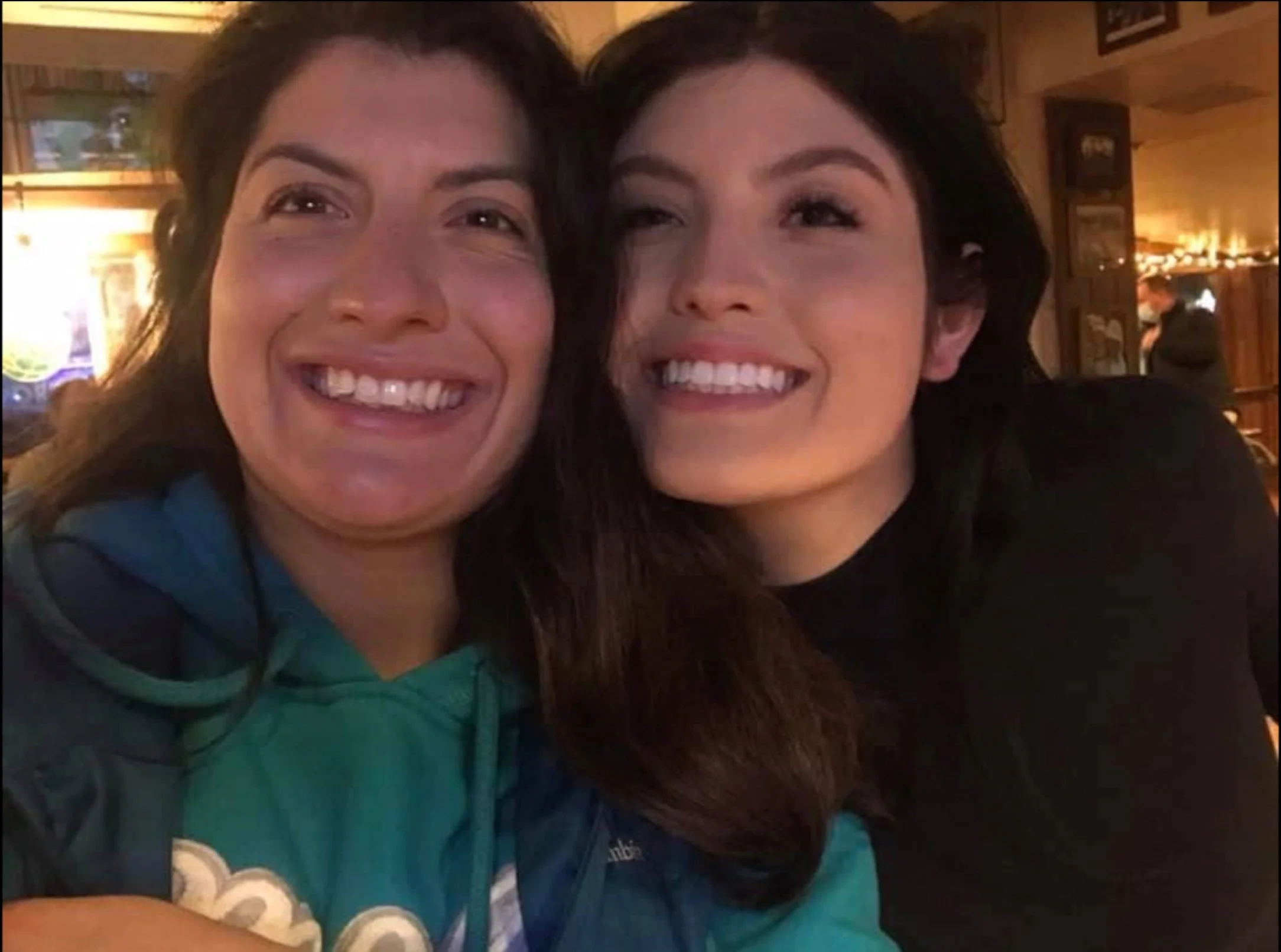 Two women smiling closely at the camera, appearing happy, in a warmly lit indoor setting.