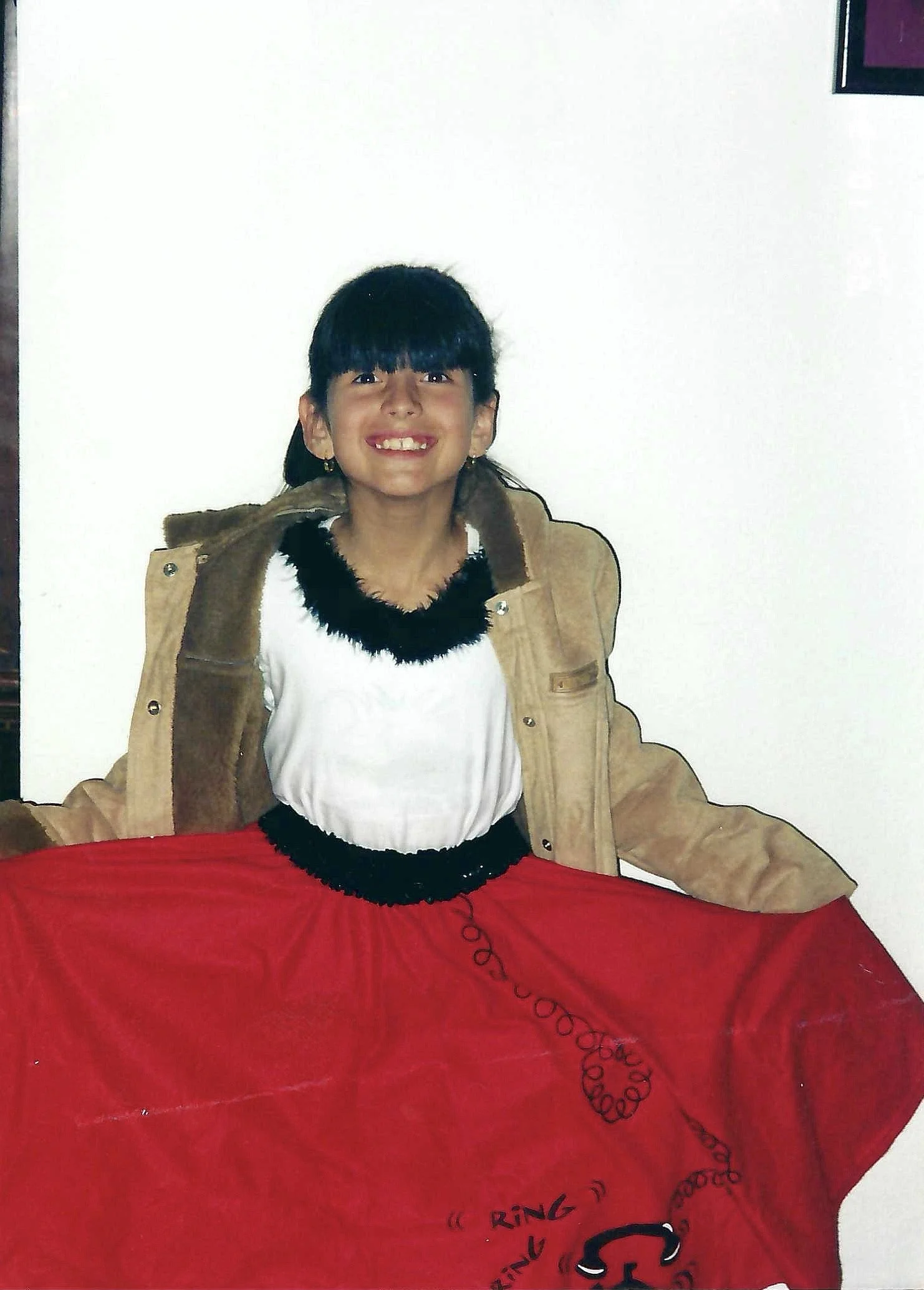 A young girl with dark hair and bangs smiling, wearing a tan jacket over a white shirt with black fuzzy collar and red skirt, sitting at a red table, with a white wall and a purple-framed picture in the background.
