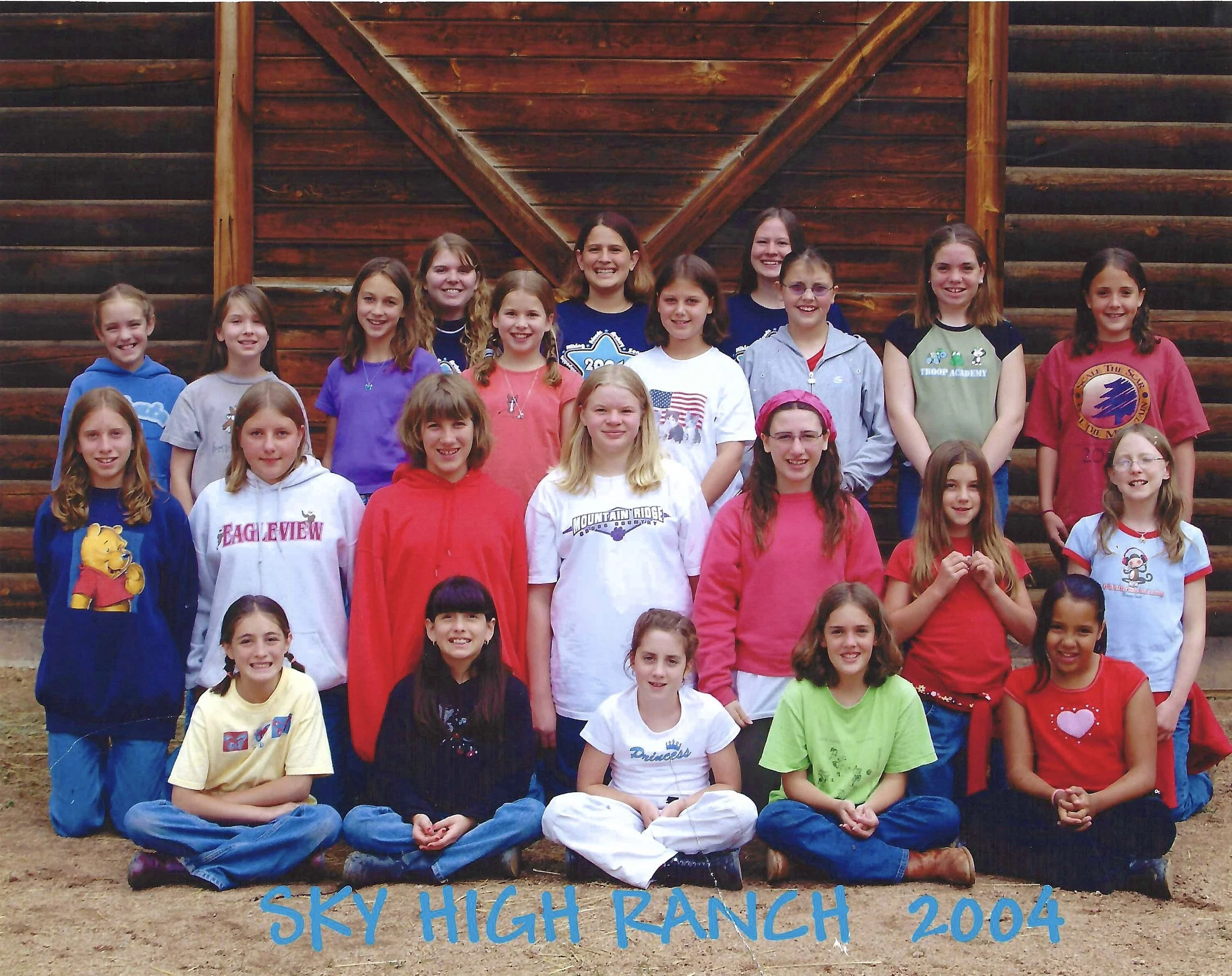 Group of children standing together in front of a wooden barn wall, some wearing colorful casual clothing and smiling.