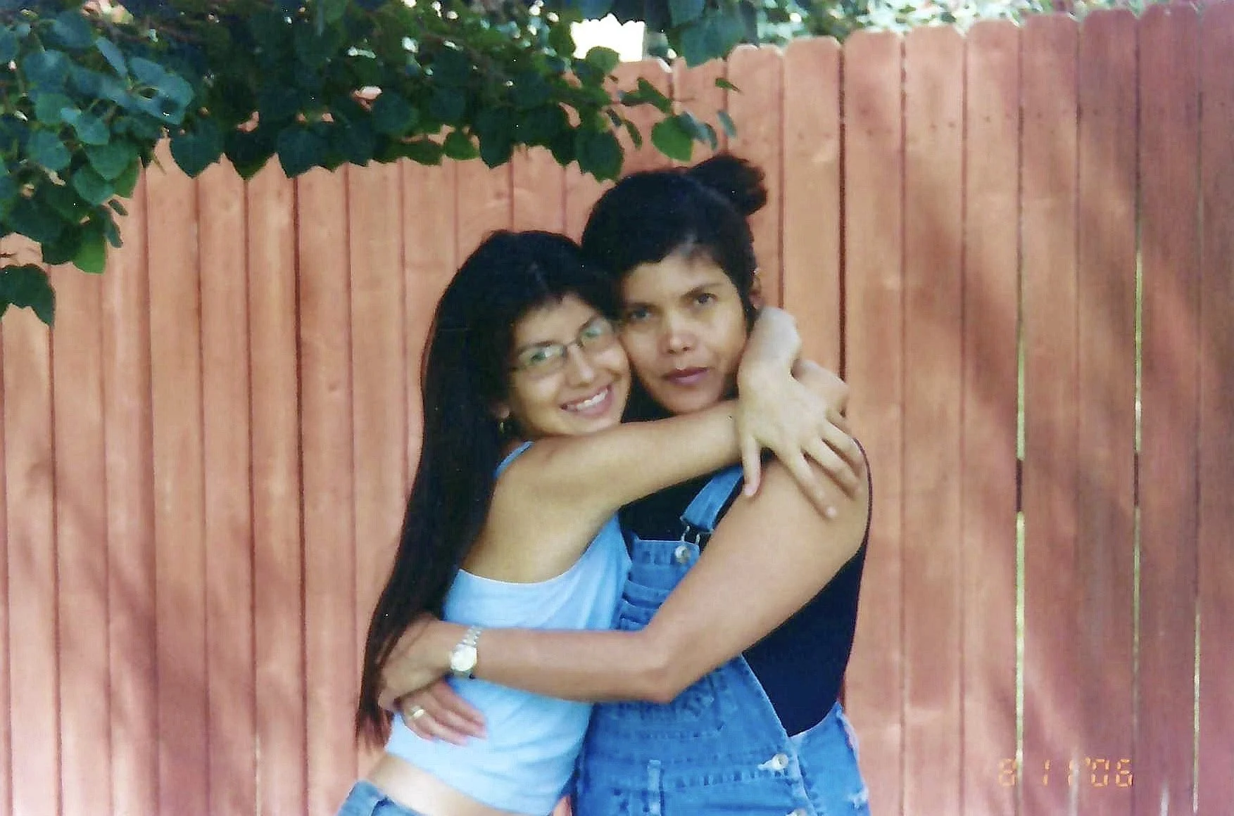 Two women hug in front of a wooden fence, one smiling and wearing glasses, the other with dark hair in a bun and wearing overalls.