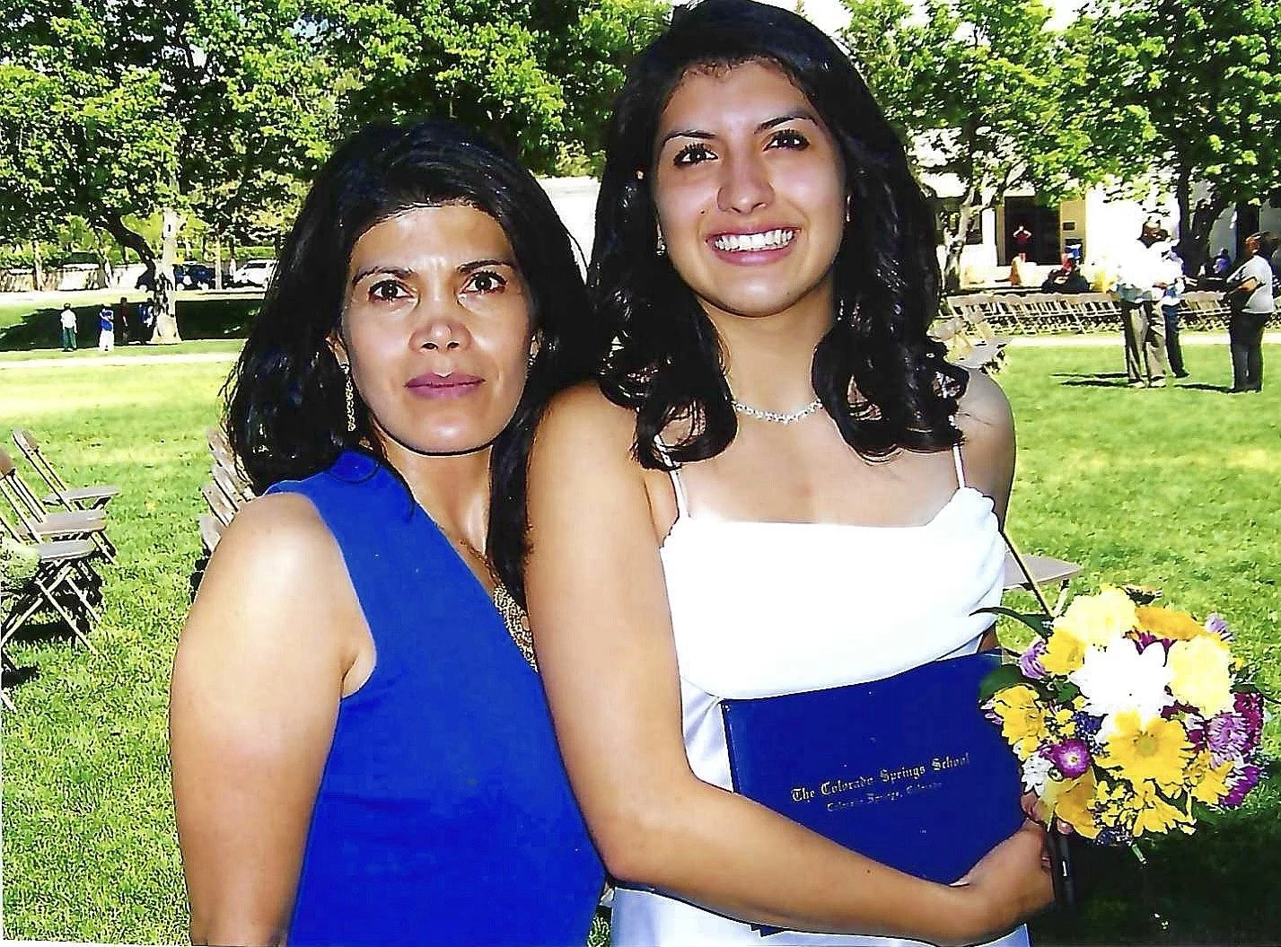 Two women outside at a graduation ceremony, one holding a bouquet of flowers and a diploma. The woman on the right is wearing a white dress, and the woman on the left is dressed in a blue dress.