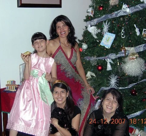 A woman and three girls celebrating Christmas in front of a decorated Christmas tree. The woman is standing behind the girls, one of whom is holding a piece of cake. The girls are sitting or standing in front of her, smiling. The scene is festive wit