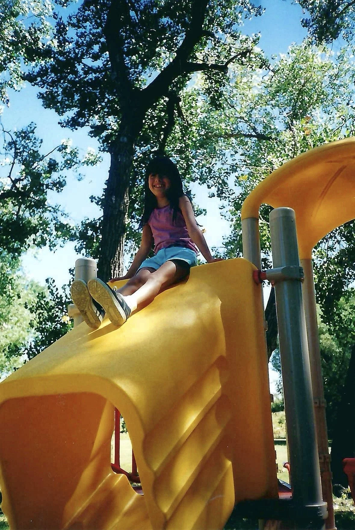 Child girl sitting at the top of yellow playground slide in a park with green trees and blue sky in the background.