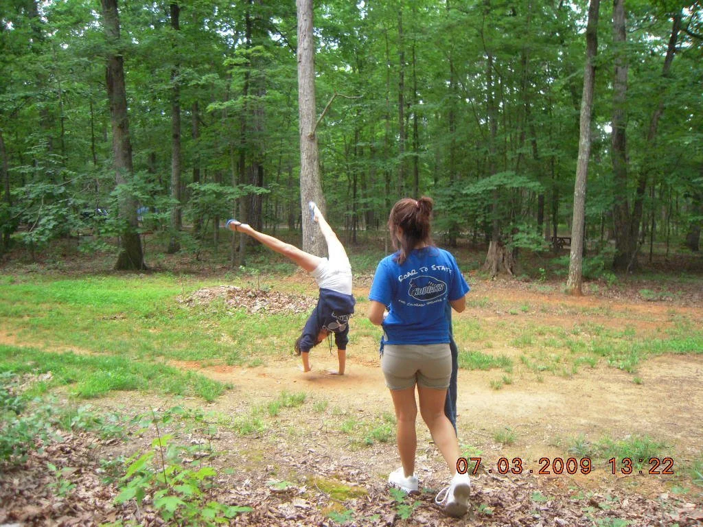 A woman wearing a blue t-shirt and shorts is watching a person handstand in a wooded area with a dirt path and green trees. The timestamp on the photo is July 3, 2009, at 1:22 PM.