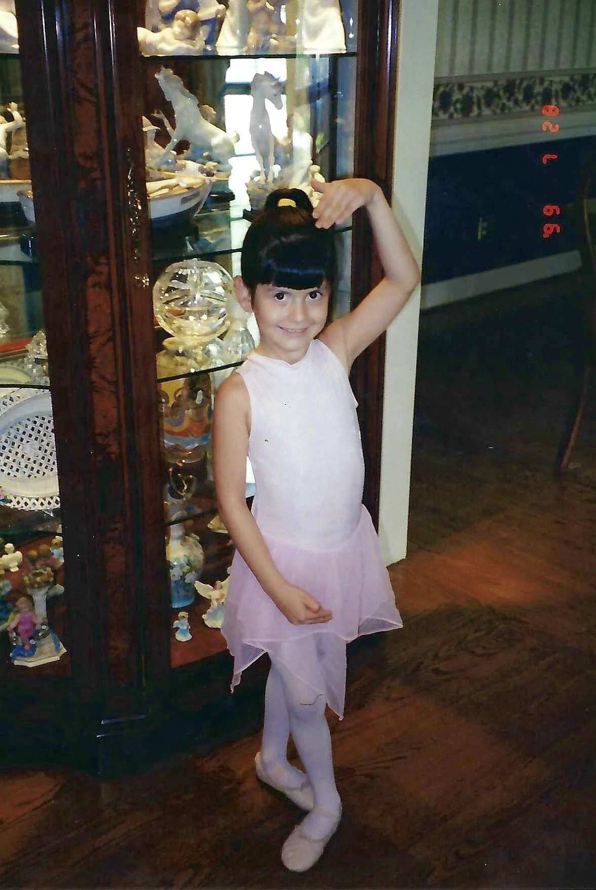A young girl dressed in a light pink ballerina outfit, with a top and a tutu skirt, standing indoors in front of a glass display cabinet filled with porcelain figurines, including horses, and various decorative items. The girl is smiling and striking