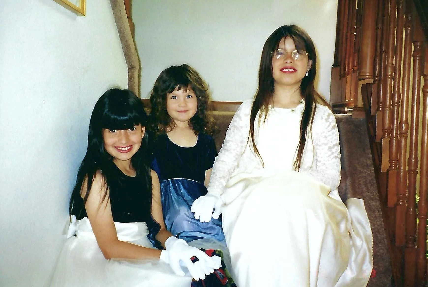 Three females sitting on a staircase. Two young girls in fancy dresses, one in a cream dress with black hair, and the other in a dark blue dress with curly brown hair. An adult woman in a white dress and glasses. The staircase has wooden railing and 