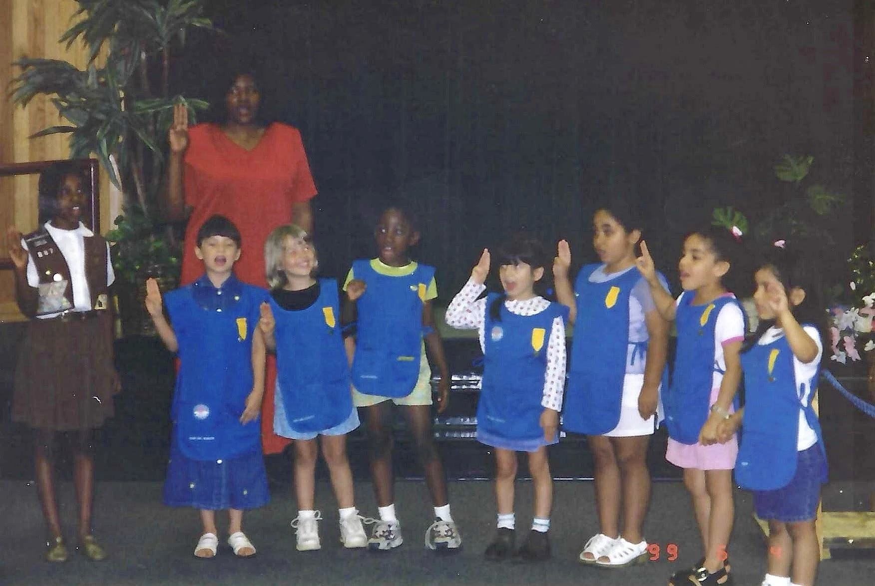 A group of children, mostly girls, standing in a line inside a room with a black background, each wearing blue vests with a yellow patch on the front, and some raising their right hands. An adult woman in a red dress is standing behind them, and a ma